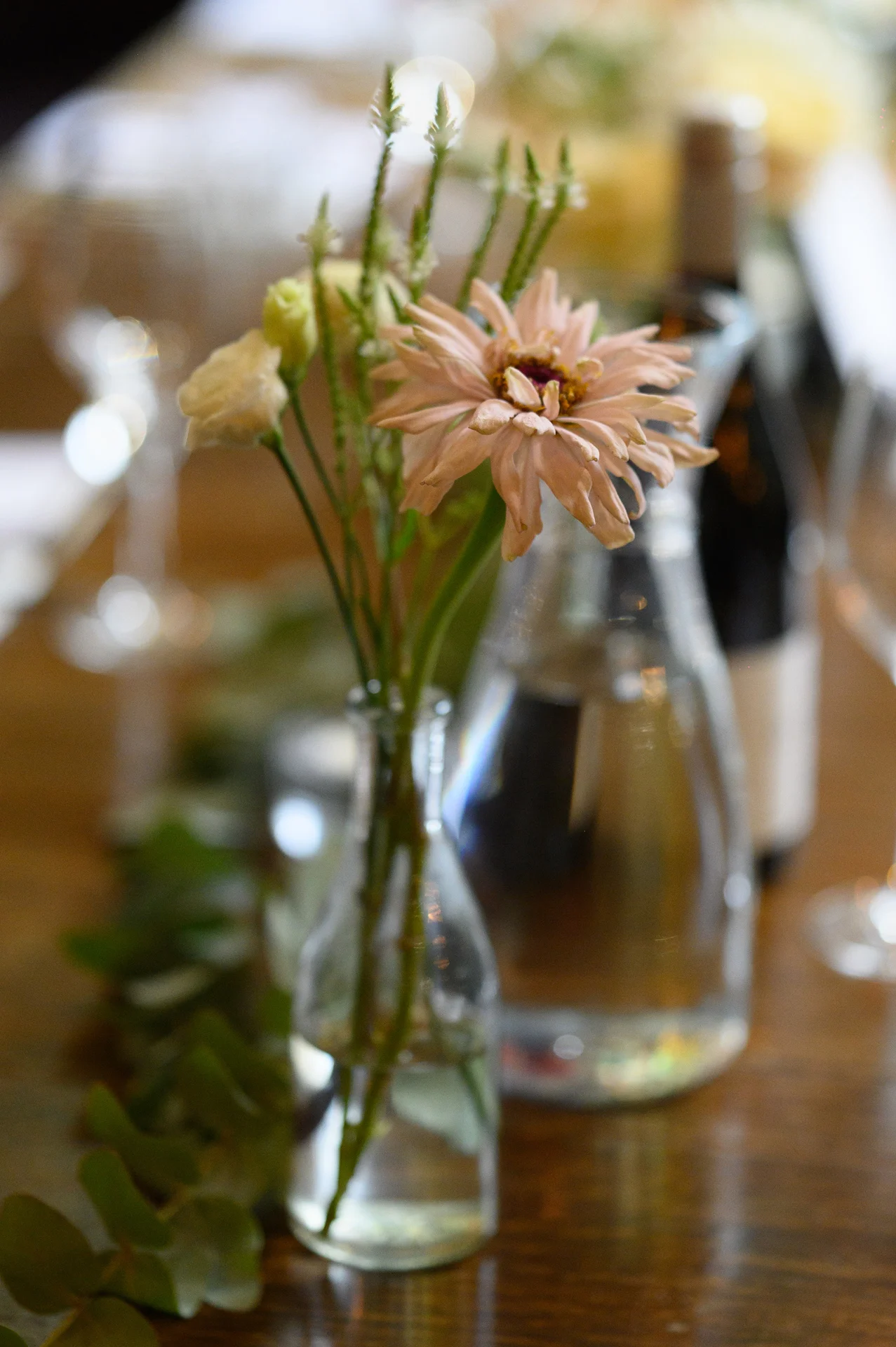 A delicate bud vase holding a single pink zinnia and wildflowers sits on a wooden banquet table among greenery runners and glassware.