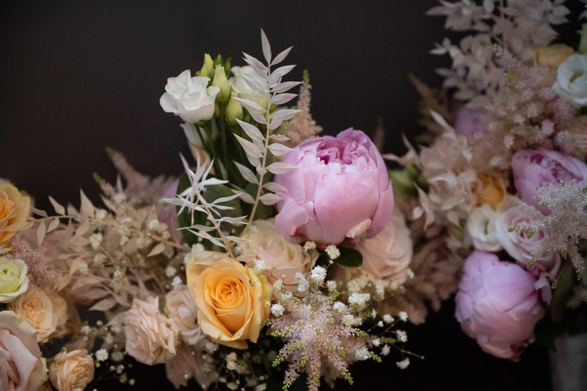 A close-up view of a stunning wedding floral arrangement featuring soft pink peonies, peach roses, and delicate astilbe against a dark background.