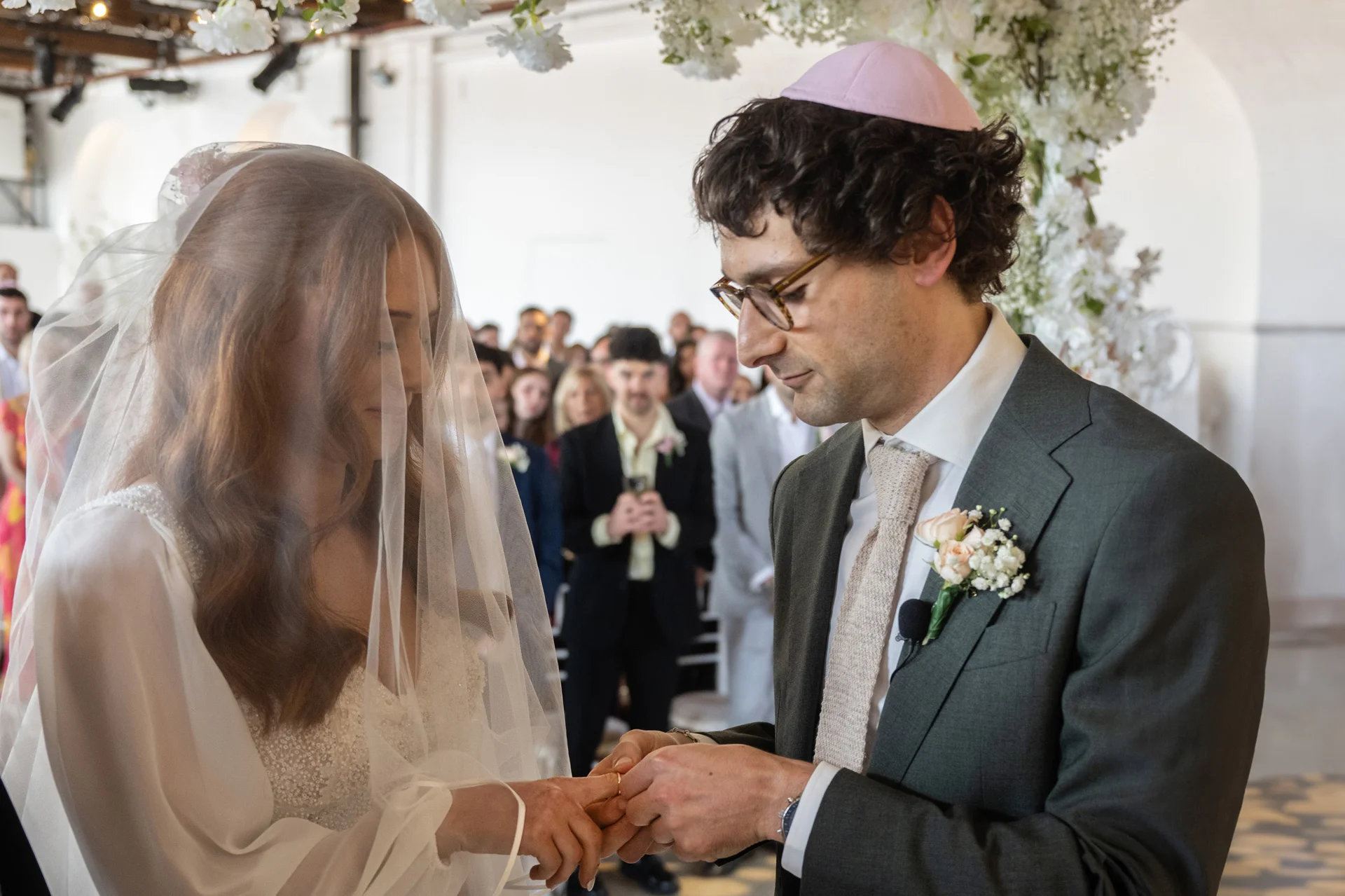 A groom gently places a wedding ring on his bride's finger during a Jewish wedding ceremony under a floral arch.