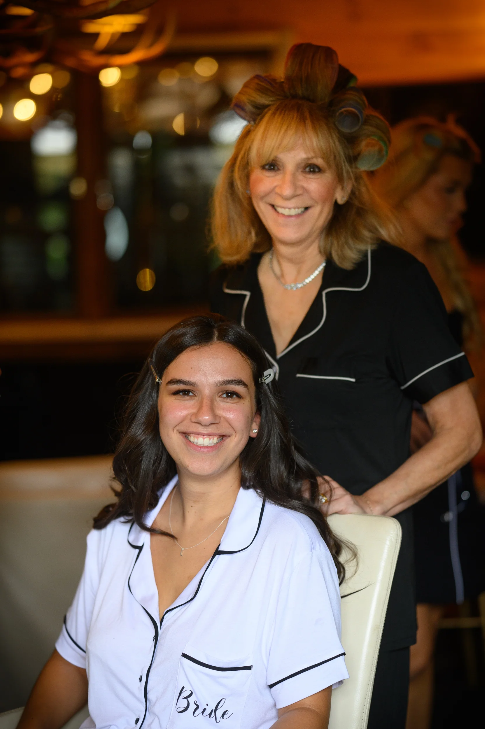 A smiling bride with dark hair wearing white pyjamas sits next to her mother who has hair rollers and is wearing black pyjamas during morning bridal prep.