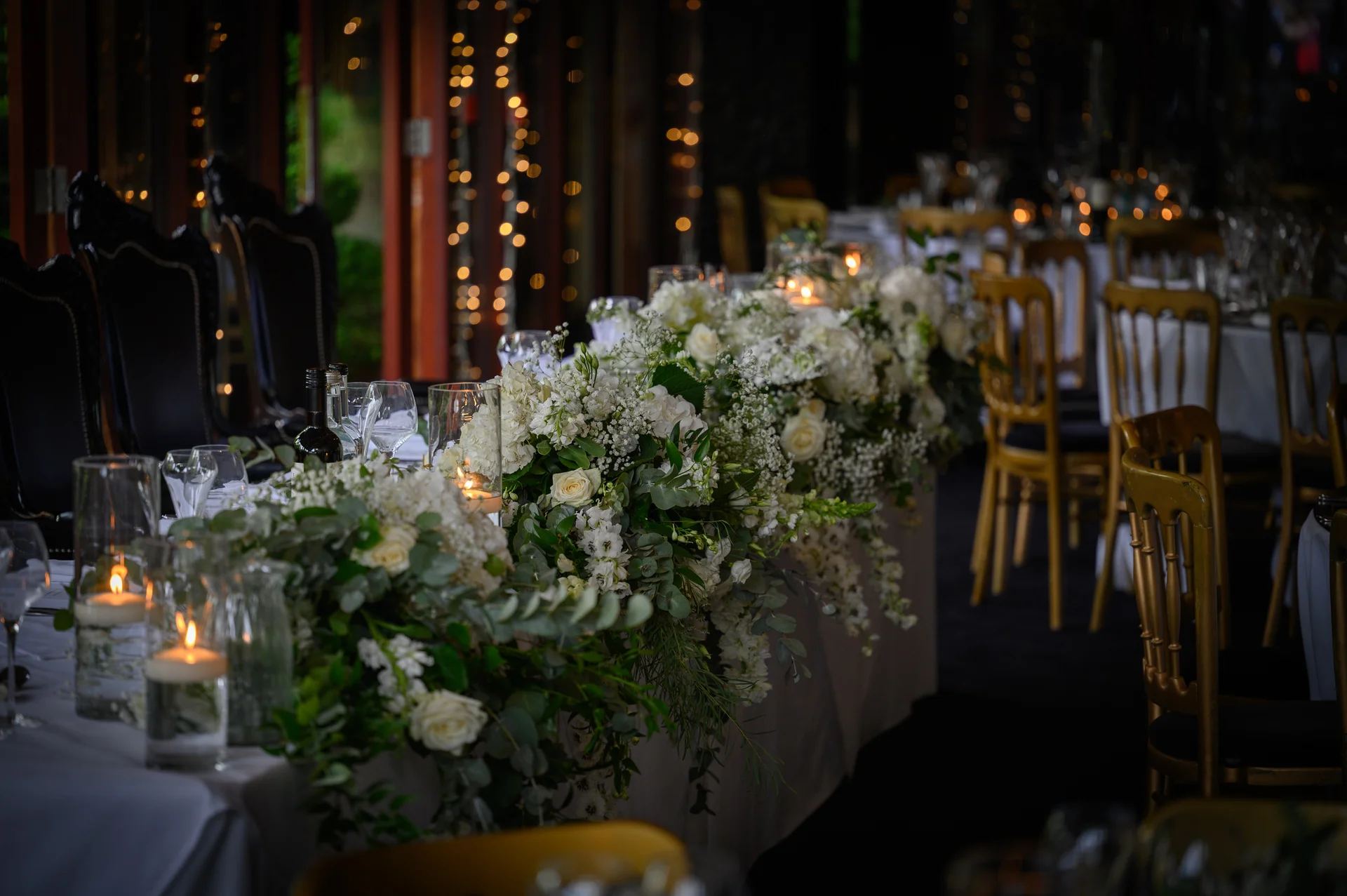 A dramatically lit wedding reception table features a lush white floral runner with roses and candlelight glowing softly along the length.
