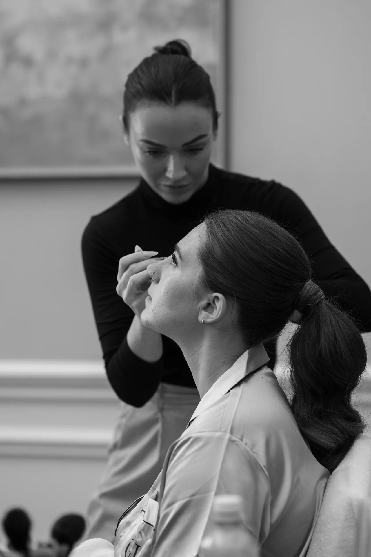 A professional makeup artist meticulously applies finishing touches to a bride wearing a robe during her morning wedding preparations.