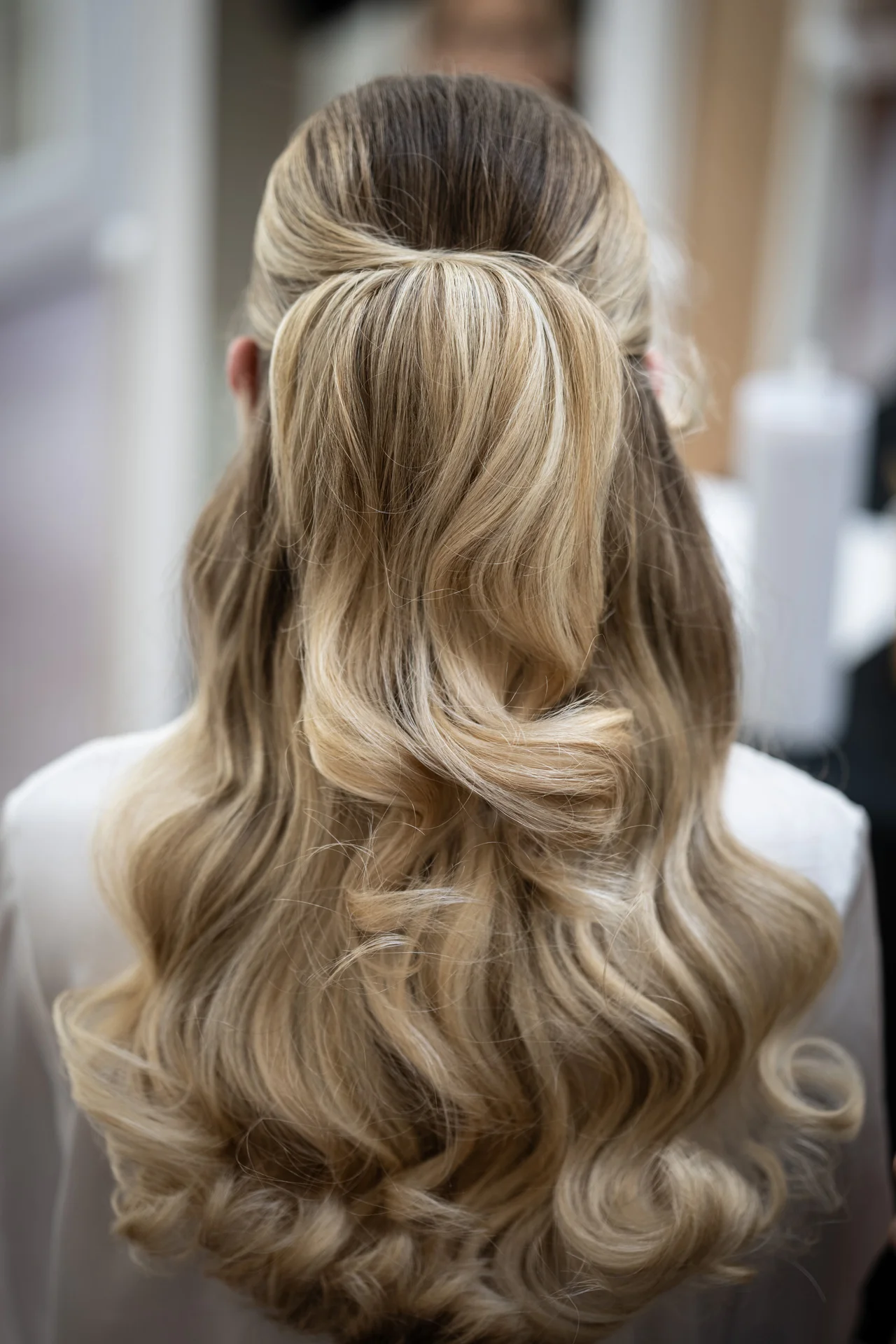 A close-up back view of a bride with blonde highlighted hair styled in a half-up half-down look with soft cascading waves during wedding morning preparations.