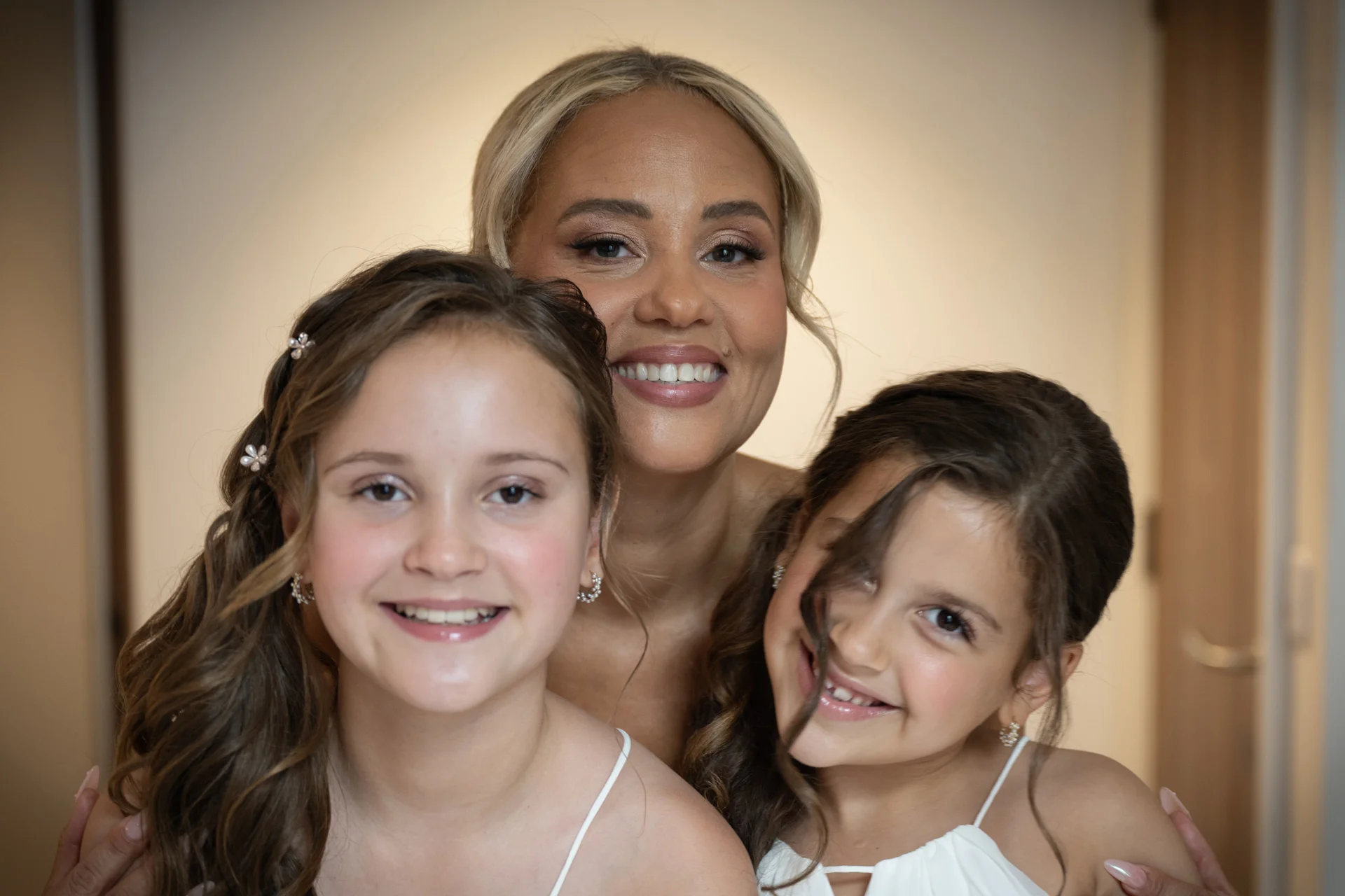 A smiling bride poses affectionately with two young junior bridesmaids styled in matching white dresses and half-up wavy hairstyles.