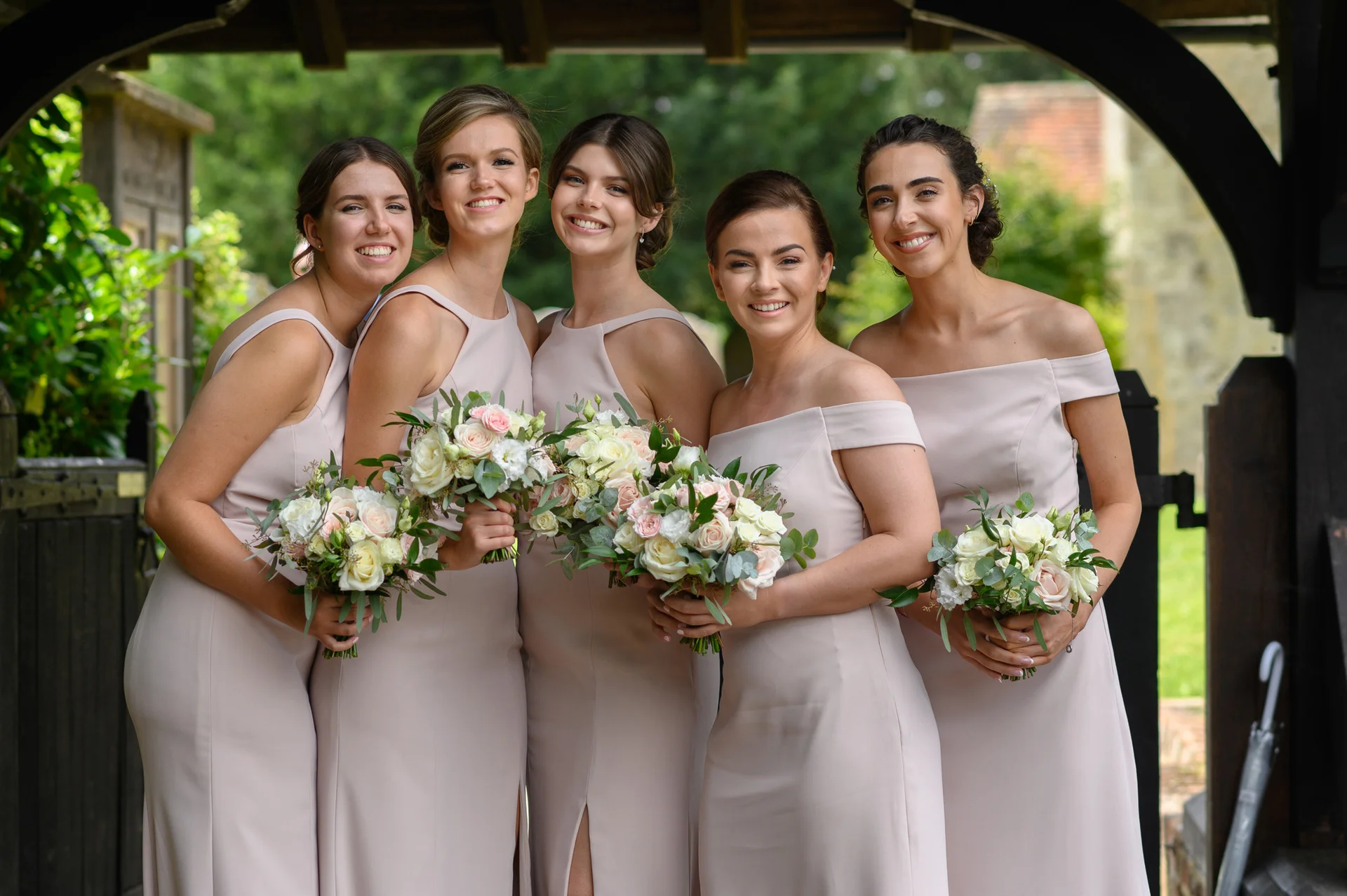Five smiling bridesmaids pose together under a dark wooden lychgate, holding matching hand-tied bouquets of roses and eucalyptus while wearing elegant blush pink dresses.