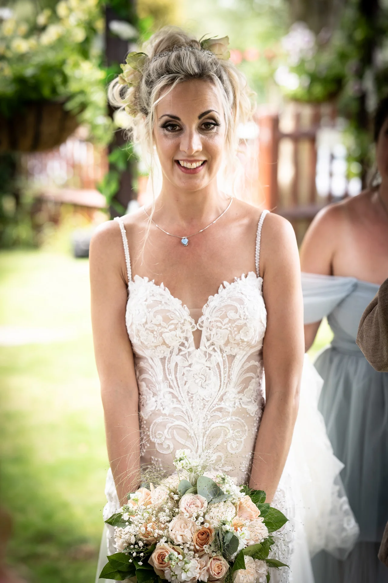 A smiling bride wearing an intricate lace gown with spaghetti straps holds a lush hand-tied floral bouquet outdoors.