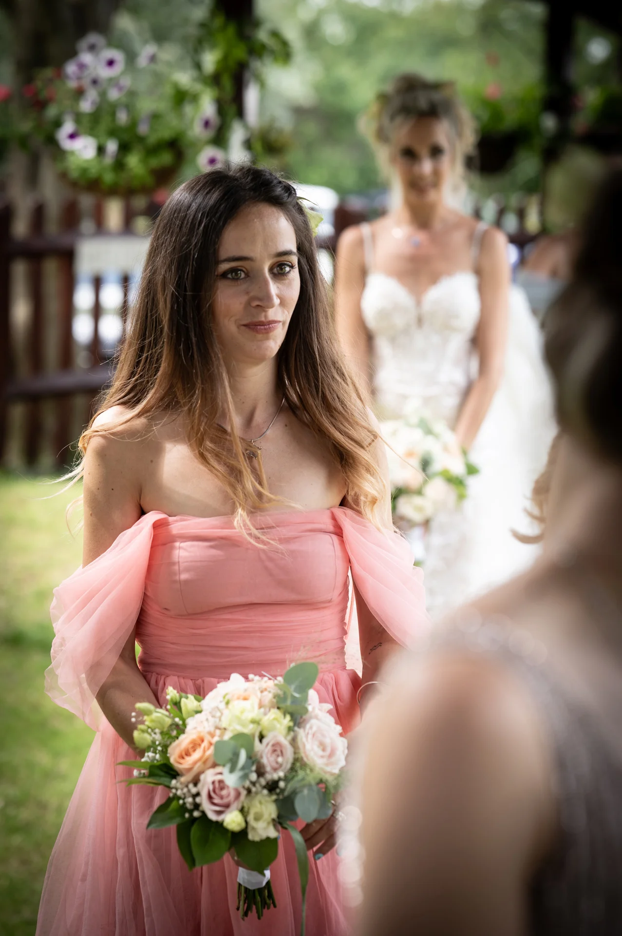 A bridesmaid in an off-the-shoulder pink dress holds a floral bouquet during an outdoor wedding ceremony, with another bridesmaid standing softly out of focus in the background.