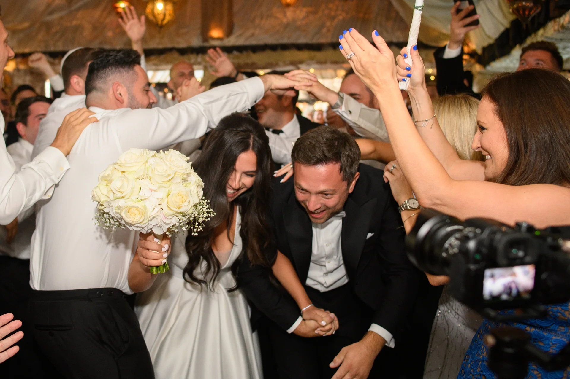 The joyful bride and groom are surrounded by cheering wedding guests on a lively, energetic dance floor during the evening reception.