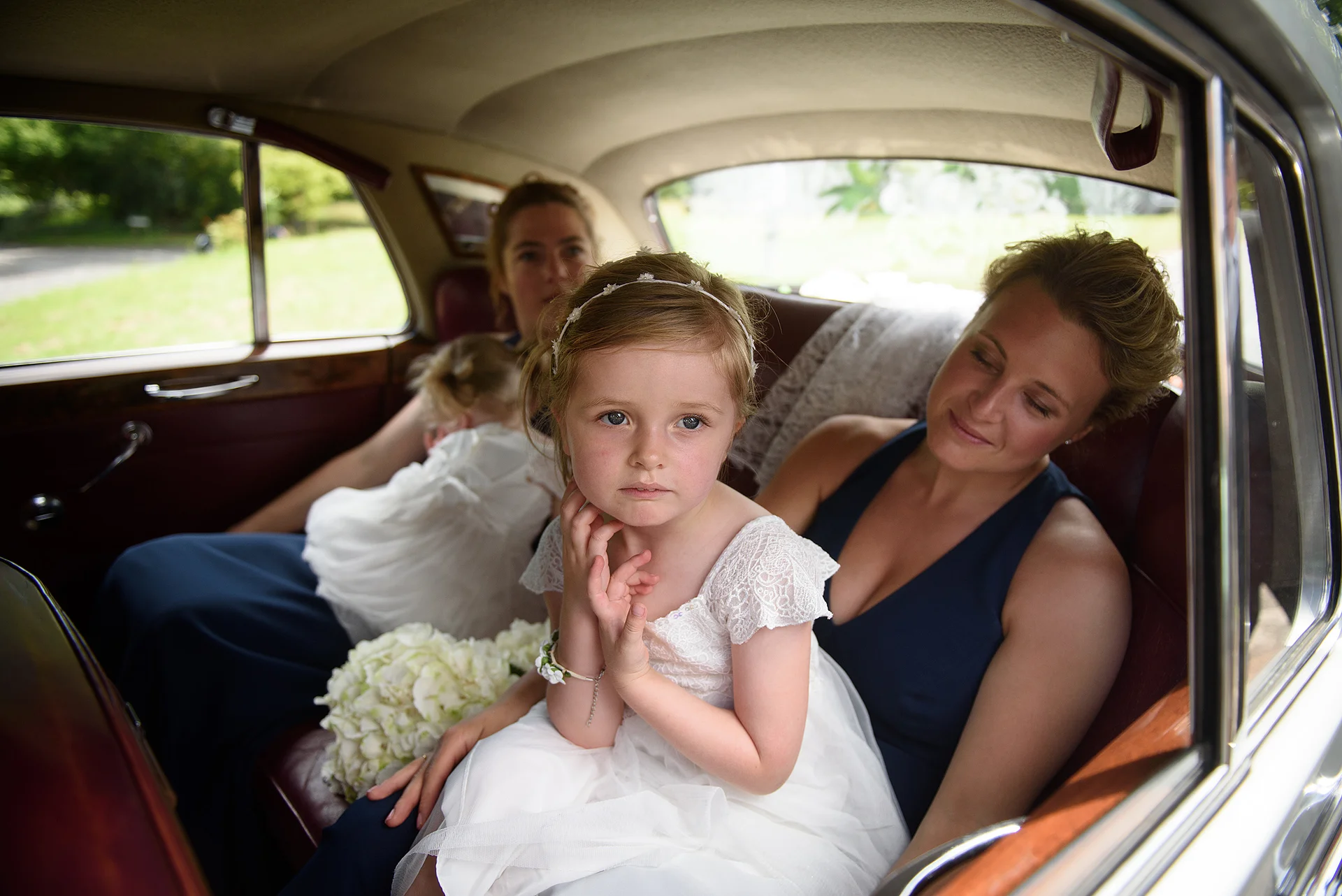 A young flower girl in a white dress sits thoughtfully in the back of a classic wedding car alongside bridesmaids.