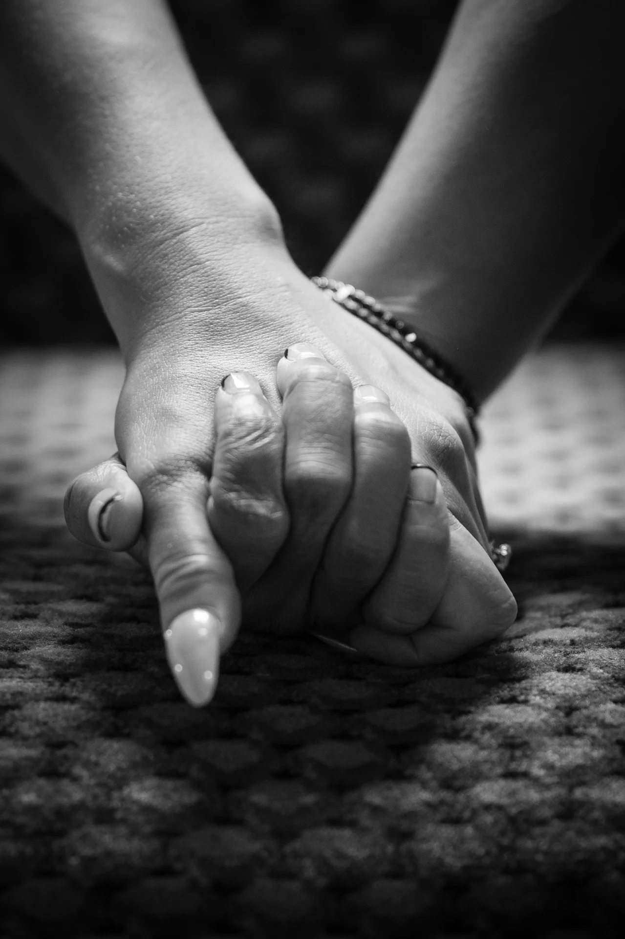 Close-up black and white shot of two people gently holding hands with intertwined fingers, capturing a quiet and intimate moment of connection.