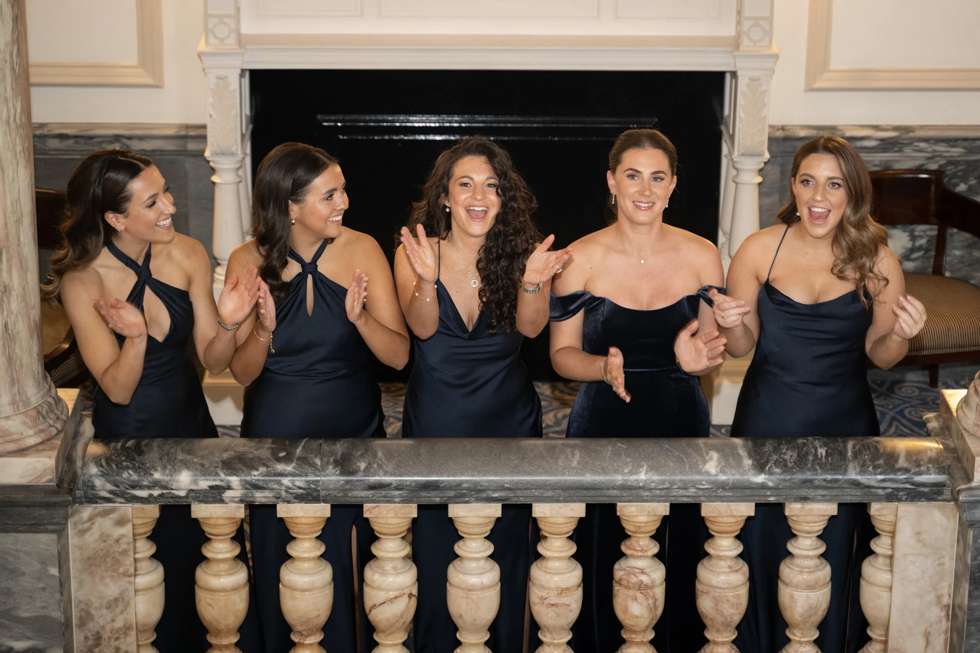 A group of five smiling bridesmaids in matching navy dresses stand behind a stone balustrade, clapping and cheering joyfully during the wedding reception.