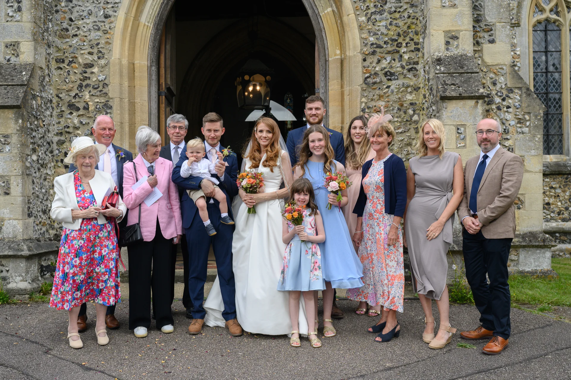 A large wedding party and family members pose together outside the stone arched doorway of a historic church after the ceremony.