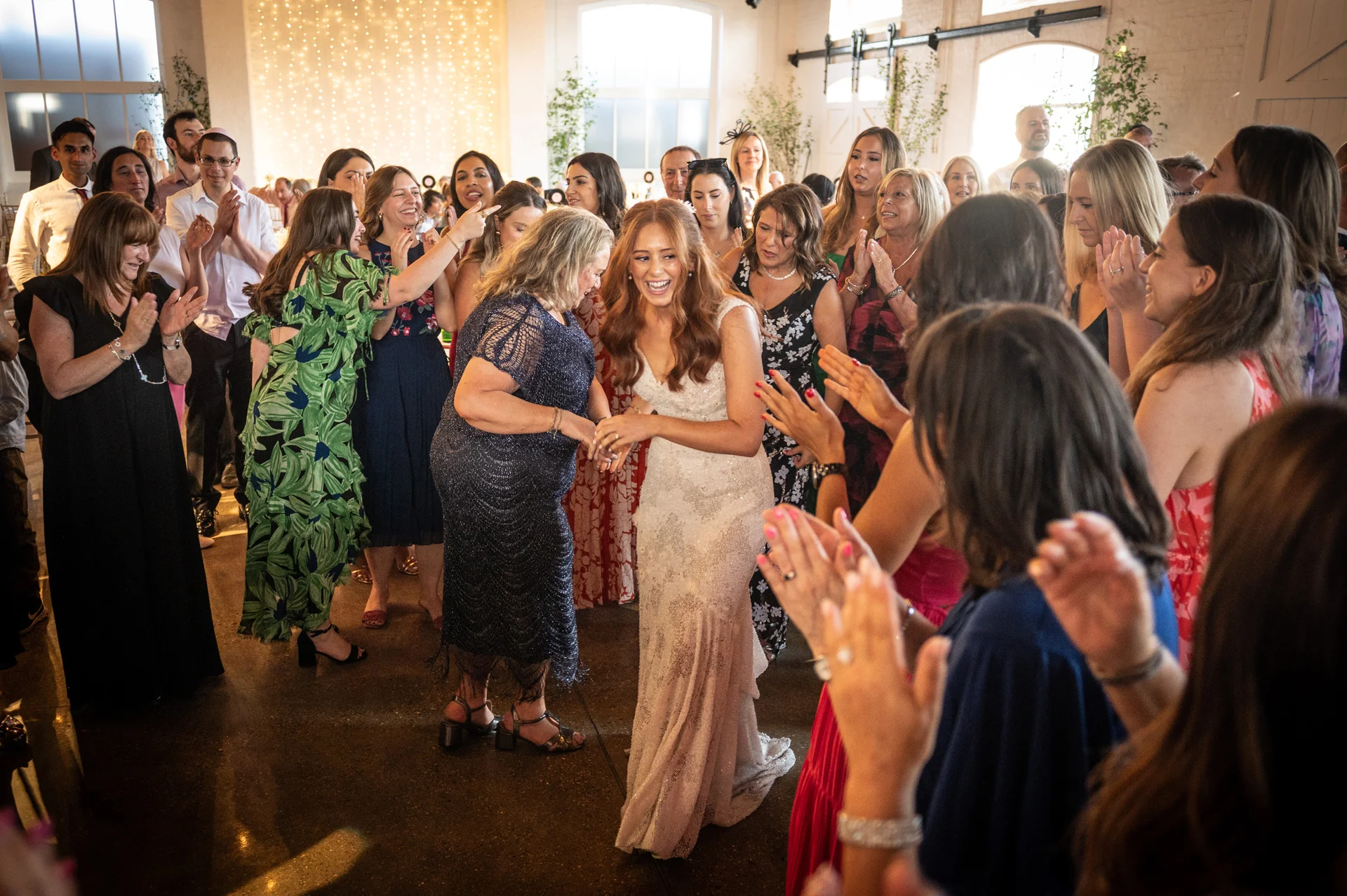 A joyful bride enters her wedding reception, laughing and smiling as guests surround her and clap enthusiastically in a bright venue.