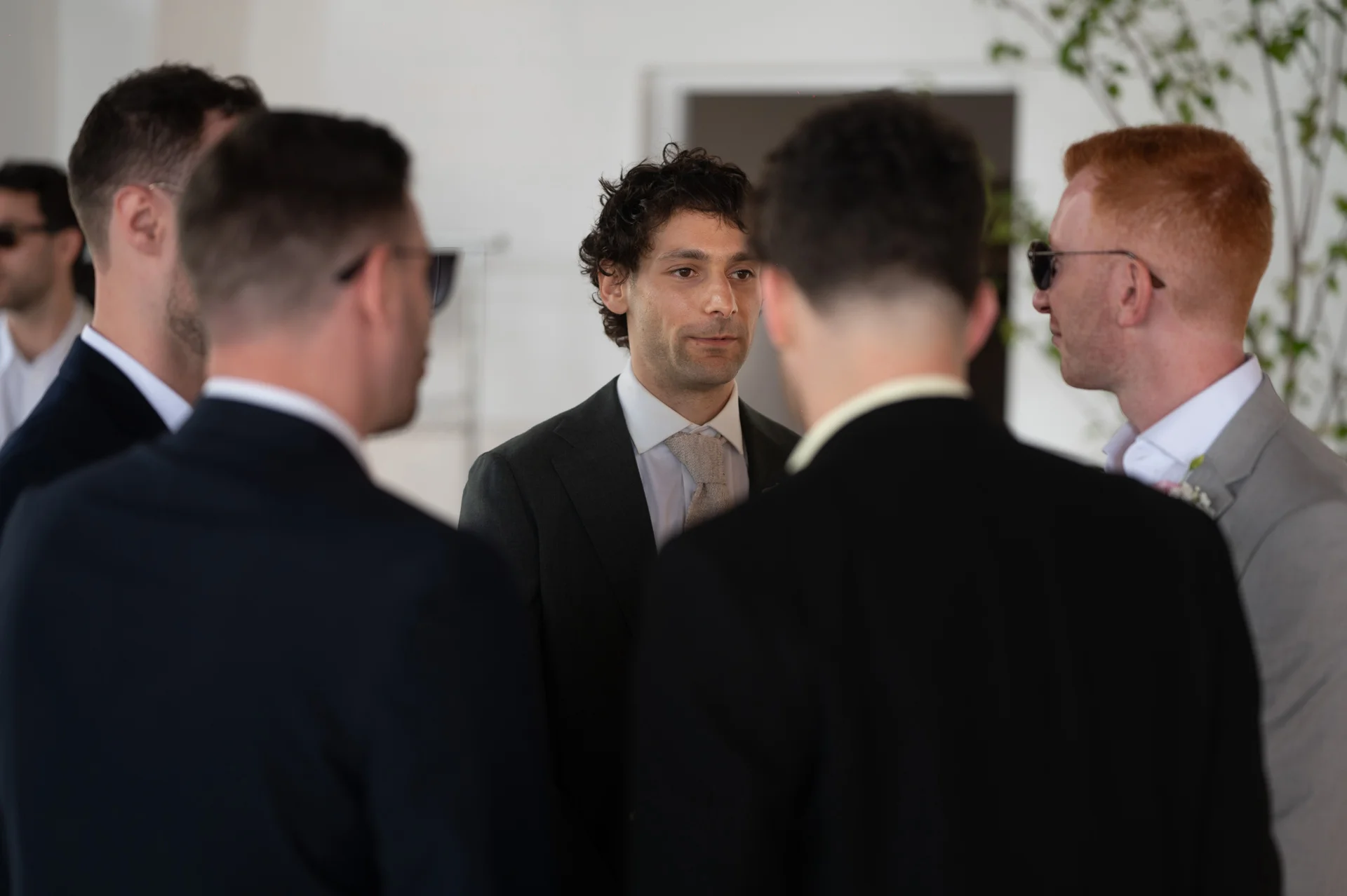 A group of smartly dressed wedding guests and groomsmen stand together indoors, chatting and sharing a lighthearted moment during the reception.