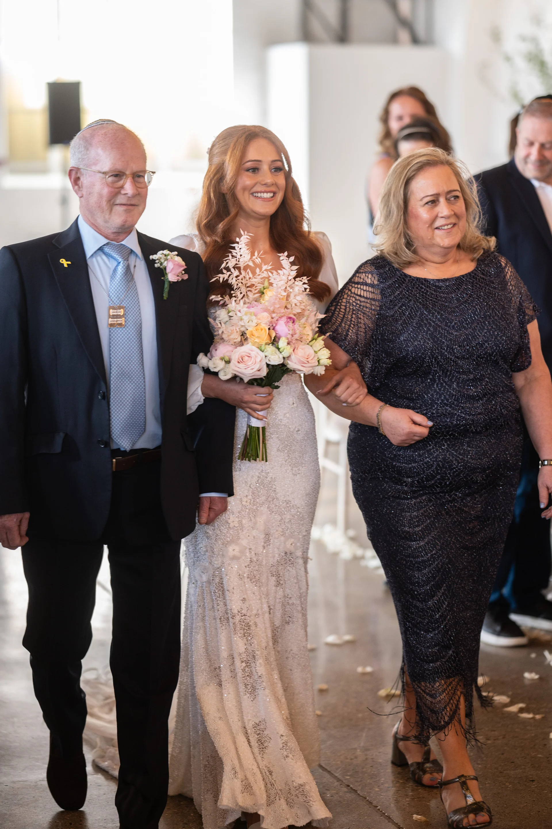 A smiling bride walking down the aisle arm in arm with her parents, holding a large pastel floral bouquet.