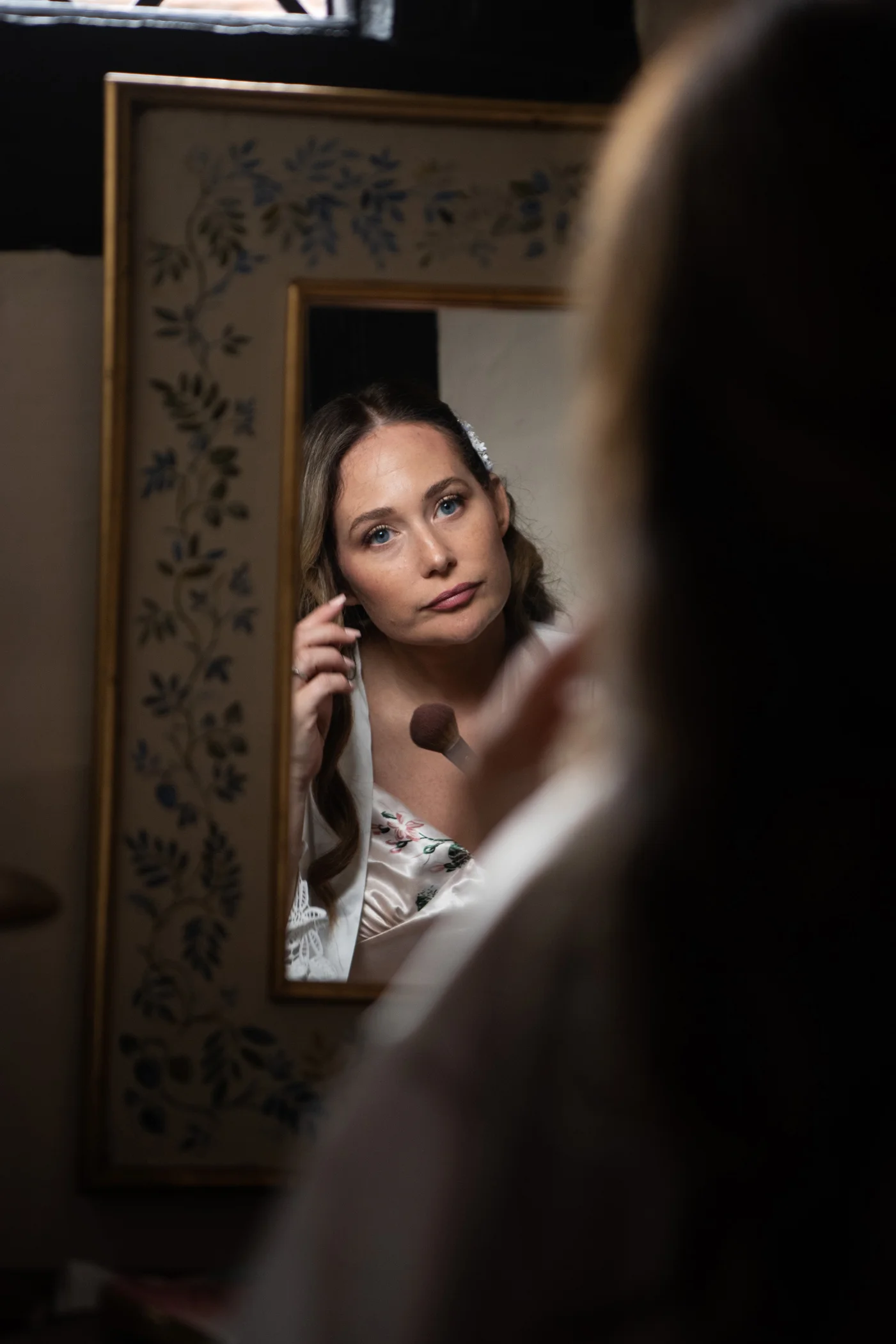 A bride looks in a decorative floral-framed mirror while applying makeup during morning bridal preparations.