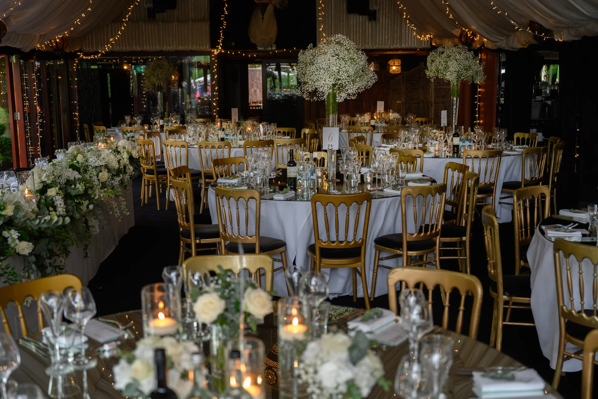 An elegantly decorated wedding marquee featuring round tables adorned with white floral centerpieces, tall candles, and gold chairs under a canopy of fairy lights.