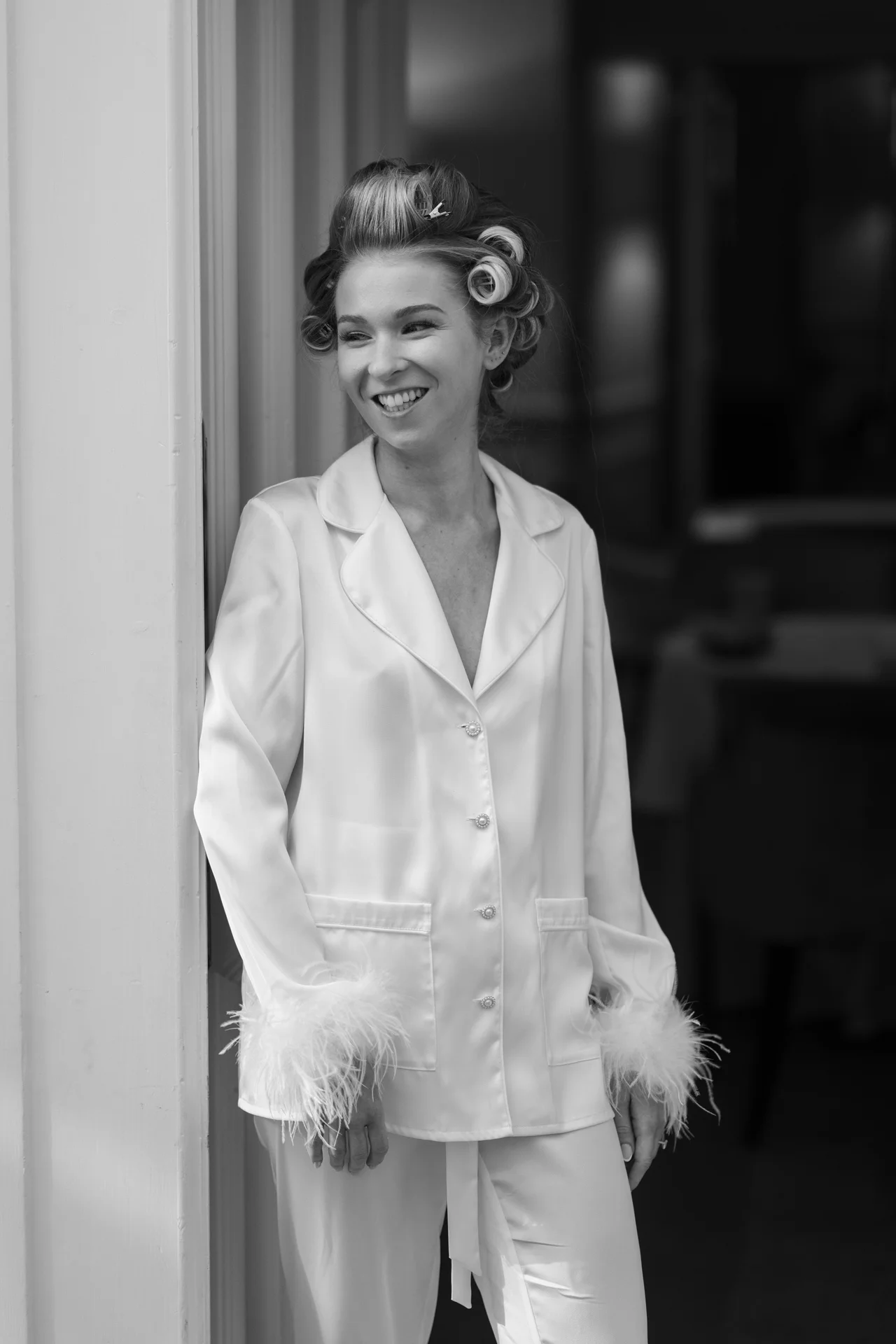 A smiling bride wearing white pyjamas with feather cuffs and hair rollers stands by a doorway during bridal preparations.
