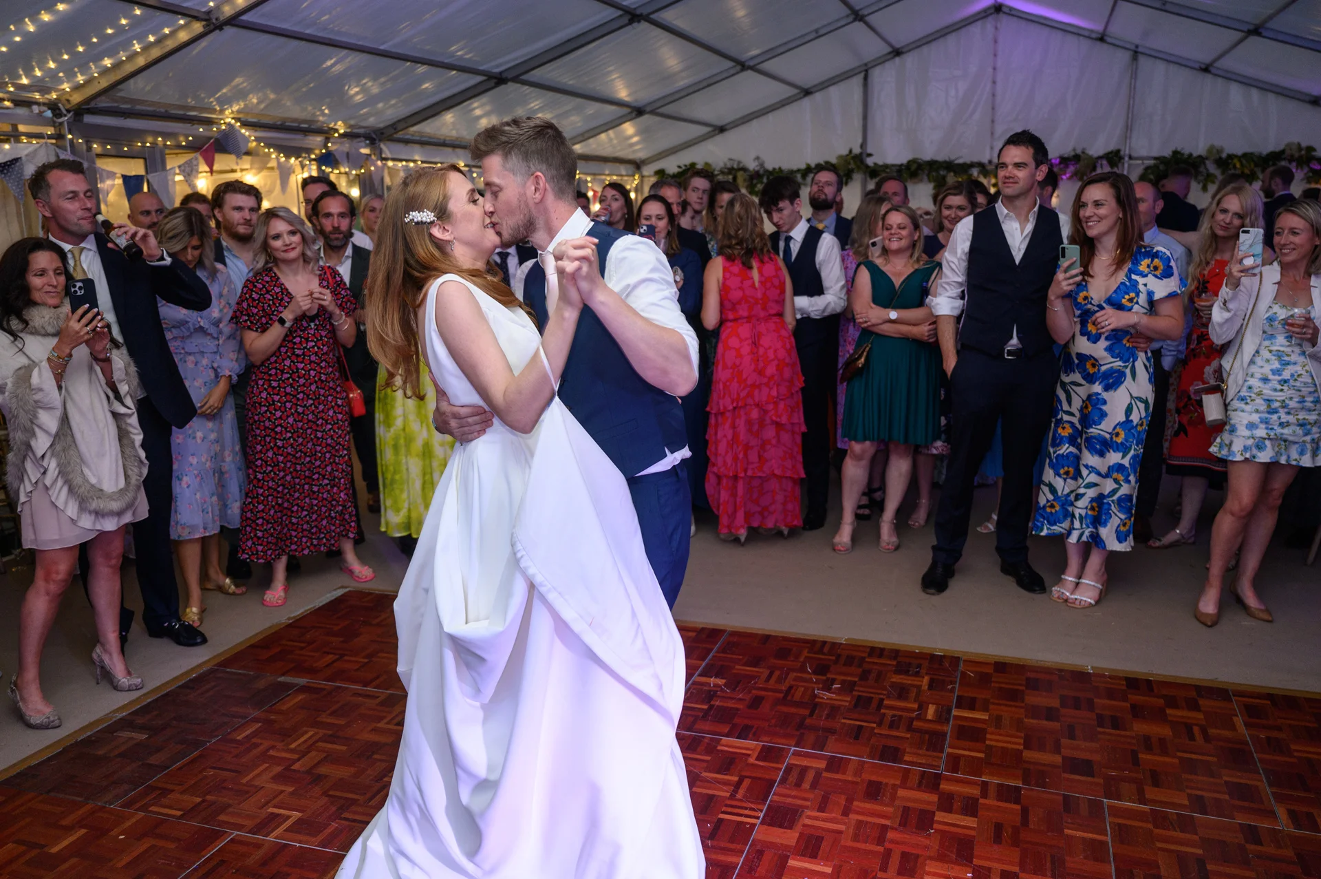 The newlywed couple share a tender first dance on a wooden dance floor inside a marquee while wedding guests watch and cheer.