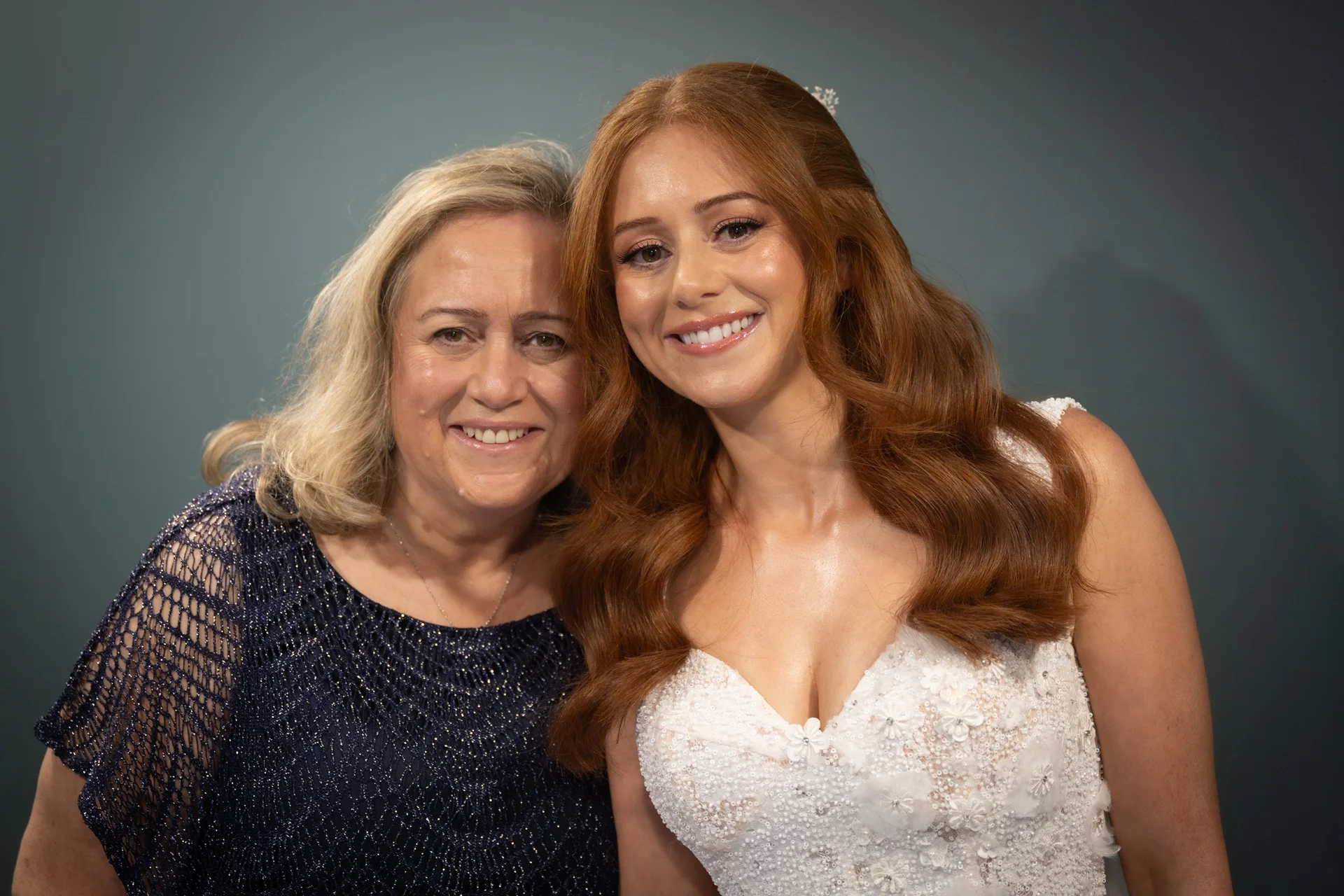 A smiling red-haired bride in a beaded floral gown poses affectionately next to her mother against a neutral dark backdrop.
