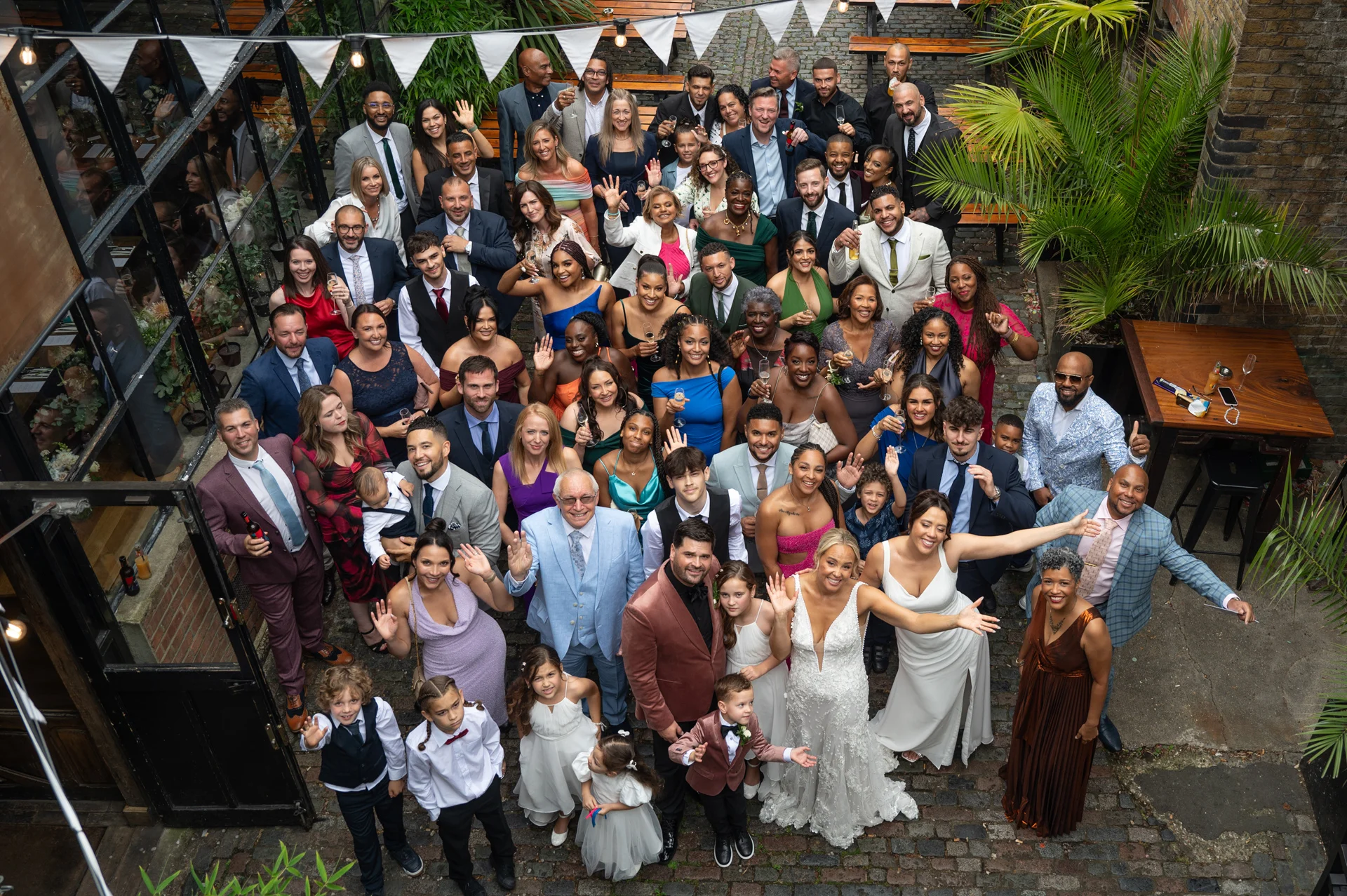 A high-angle group shot of a large wedding party and guests cheering and waving in an industrial pub courtyard decorated with bunting.