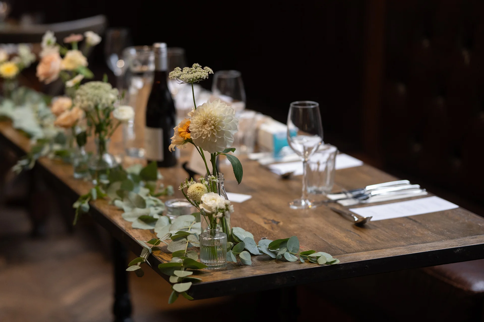 An elegantly styled long wooden banquet table features delicate bud vases with white dahlias and a cascading eucalyptus greenery runner.