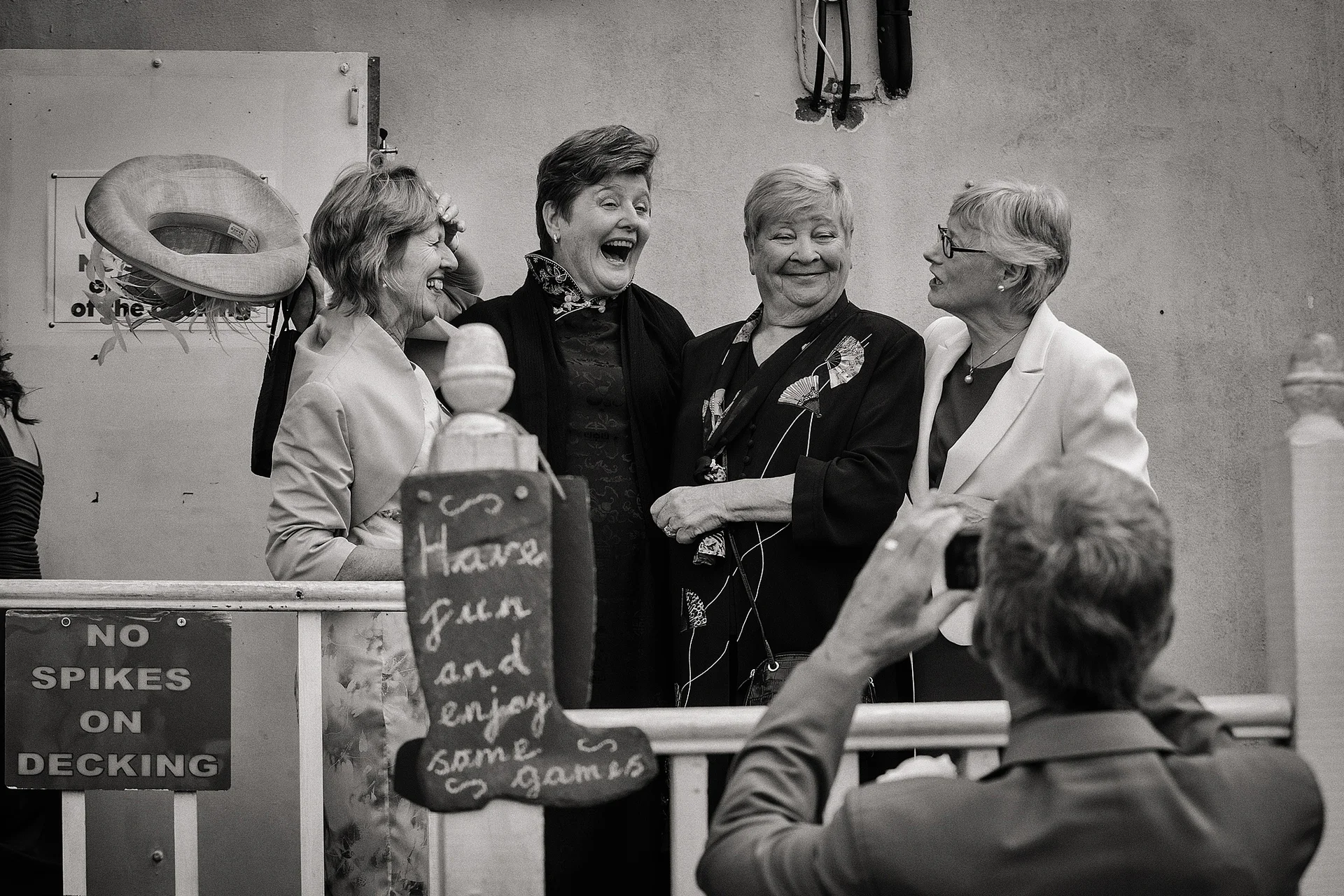 A group of older female wedding guests laughing candidly as someone takes their picture outdoors by a wooden railing and a chalkboard sign.
