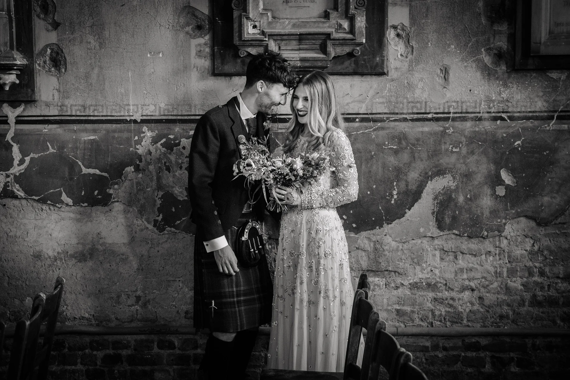 A newlywed couple in traditional Scottish highland dress and a beaded long-sleeve gown laugh together against a weathered historic wall.
