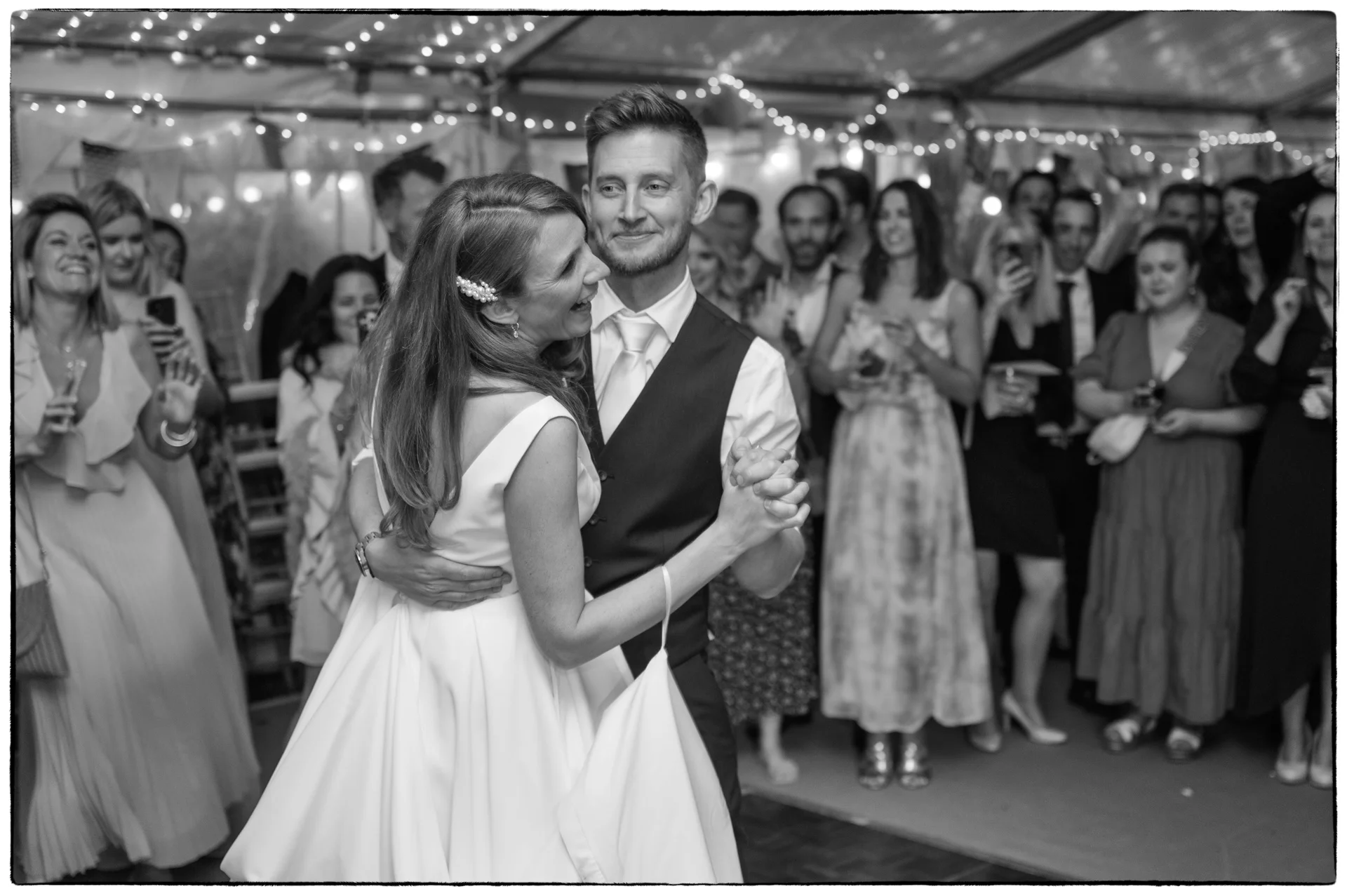A newlywed couple shares their first dance inside a marquee illuminated by fairy lights, surrounded by cheering wedding guests.