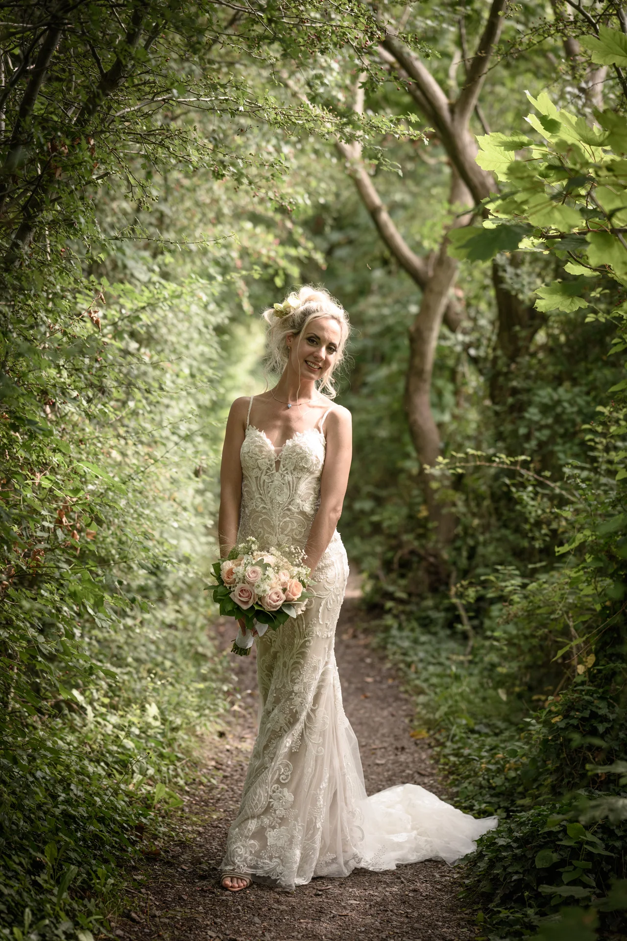A smiling bride stands on a narrow pathway surrounded by lush green trees and foliage, holding a soft pastel rose bouquet.
