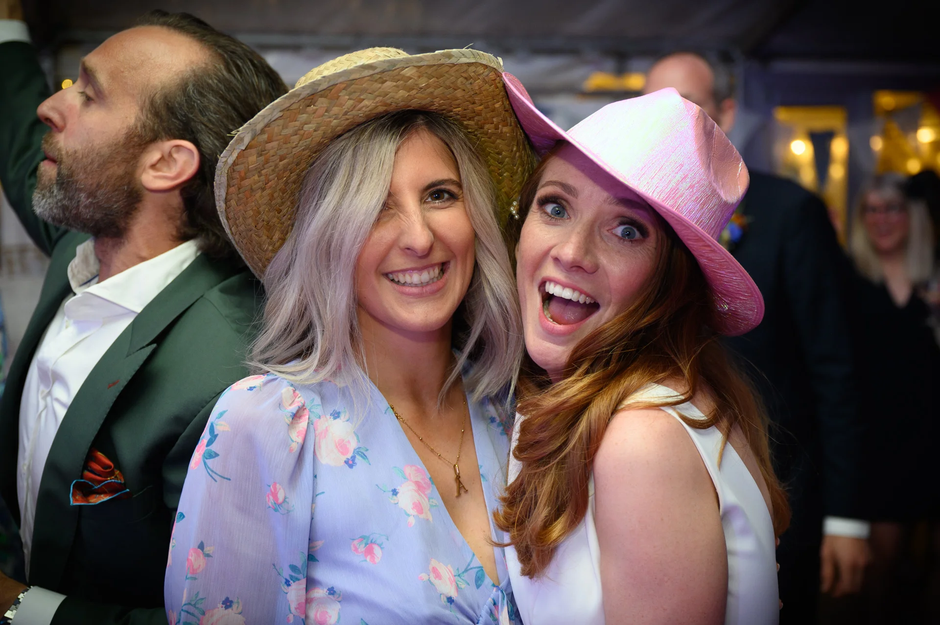 Two smiling and laughing female wedding guests wearing fun hats pose closely together at an evening reception.