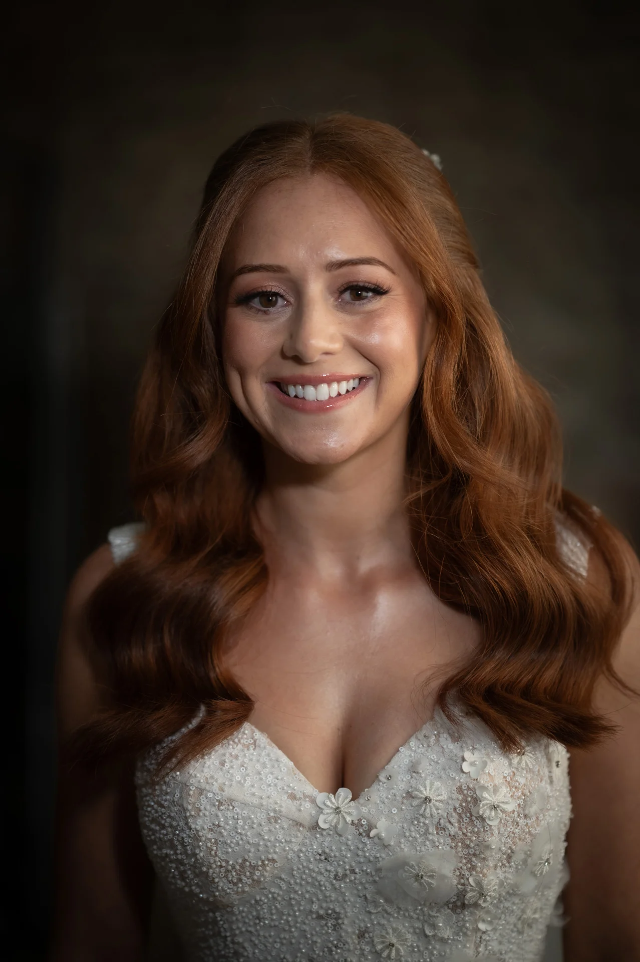 A smiling red-haired bride with soft waves and glowing makeup poses in a detailed beaded gown with a sweetheart neckline.