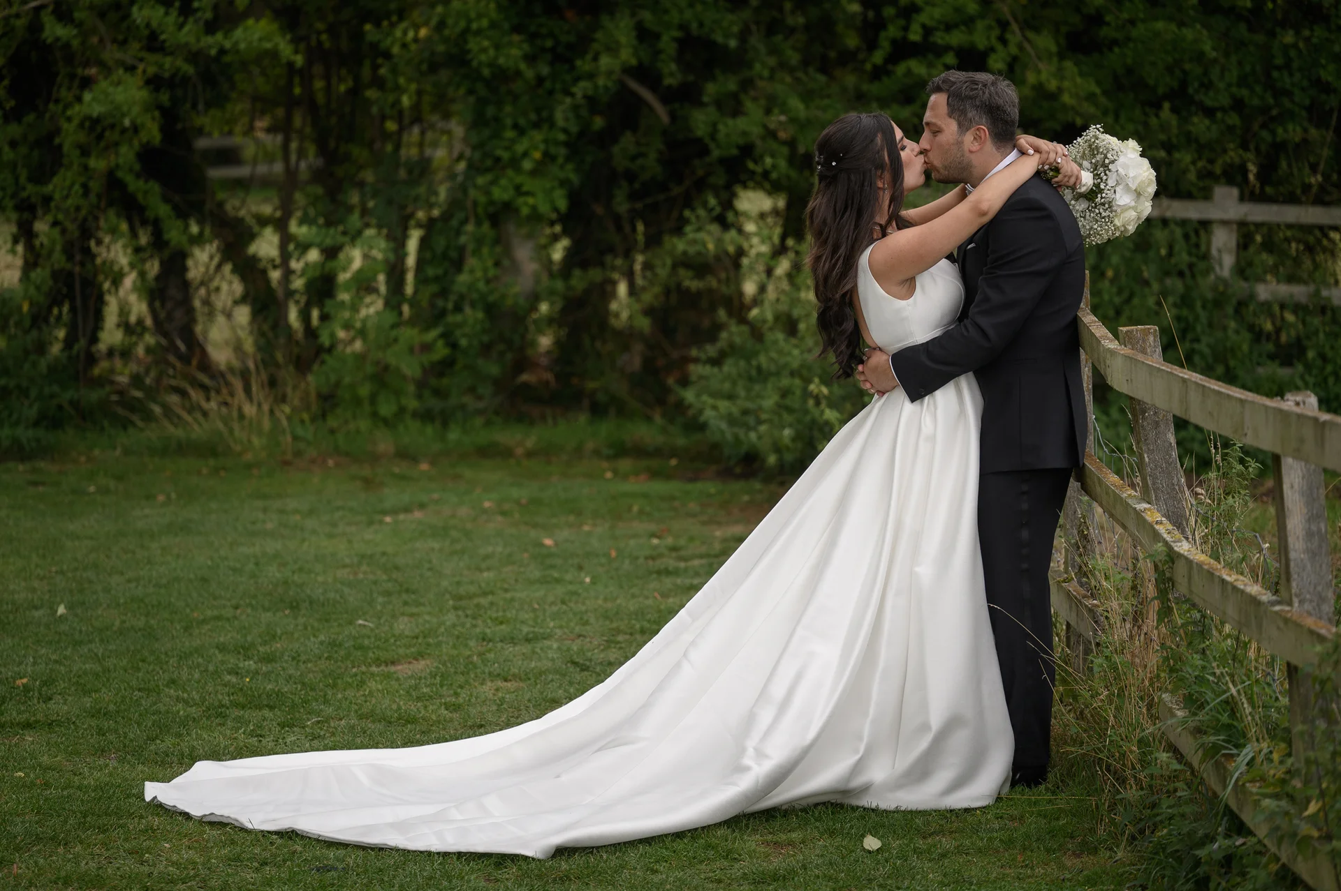 Newlywed bride and groom share a tender kiss by a rustic wooden fence in a lush green countryside setting.