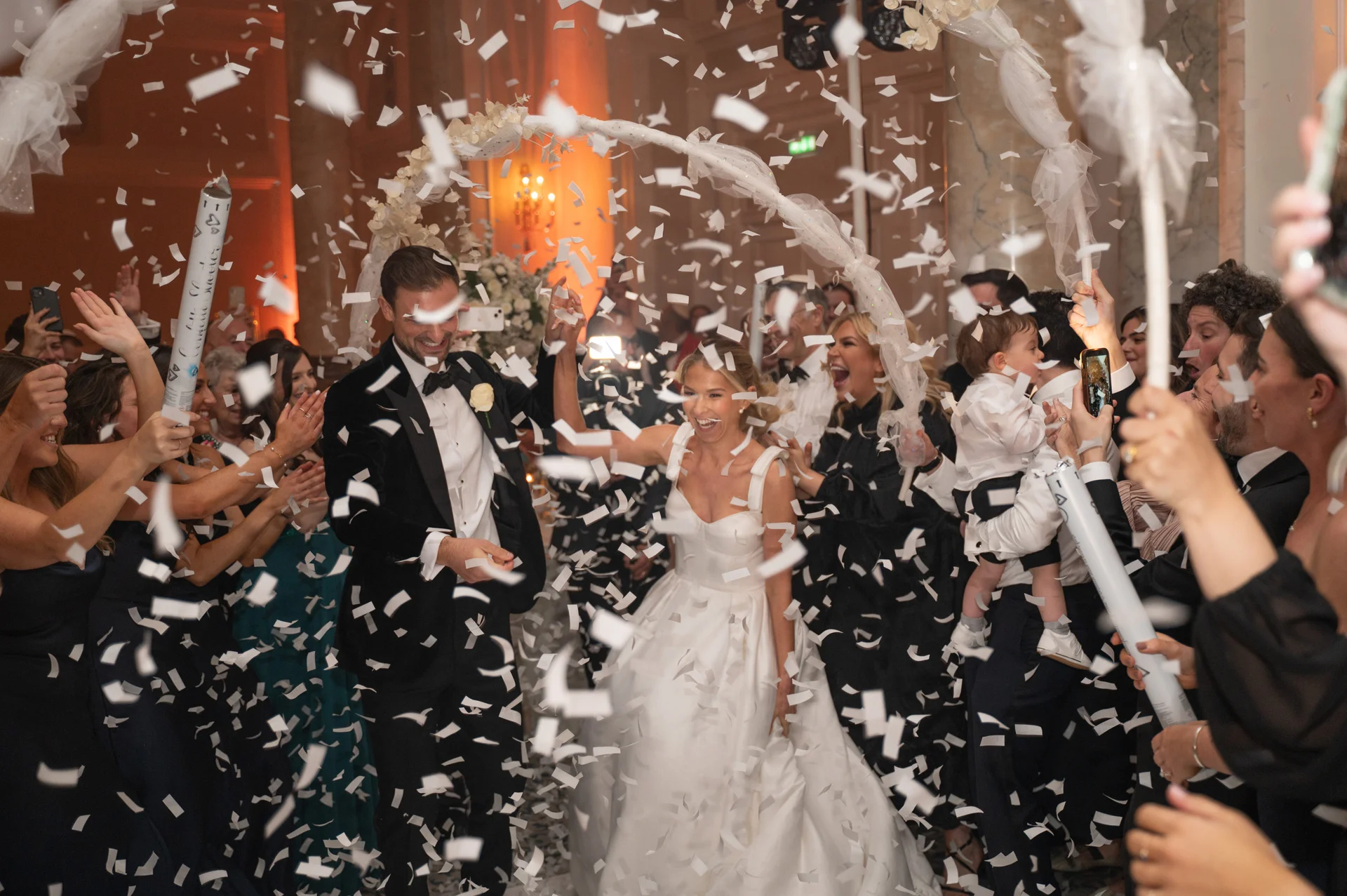 The joyful newlyweds walk hand in hand through a cheering crowd of guests while white confetti rains down around them under an arched entrance.