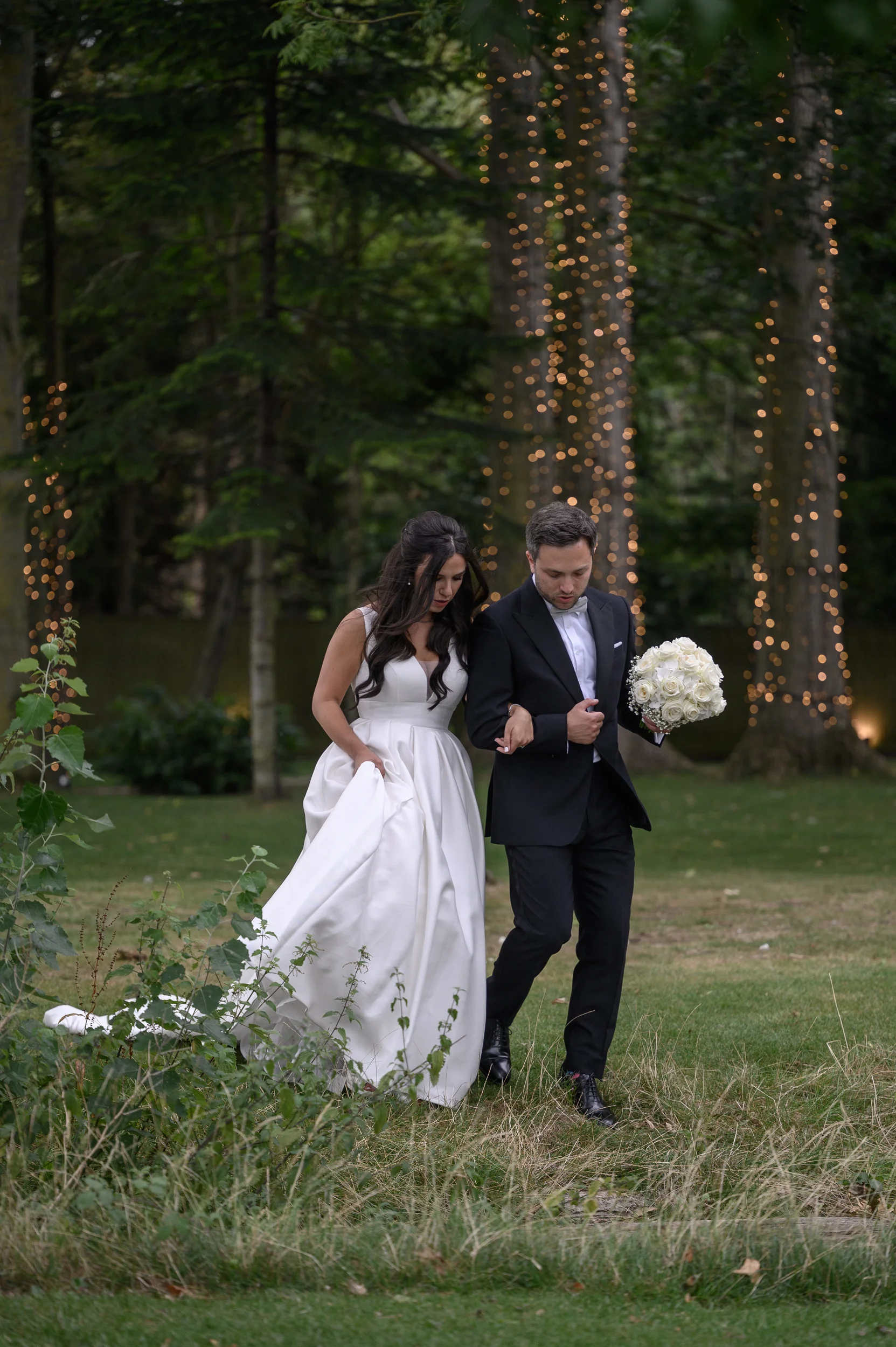 A bride and groom walk together through a woodland clearing draped with fairy lights, with the bride holding a classic white rose bouquet.