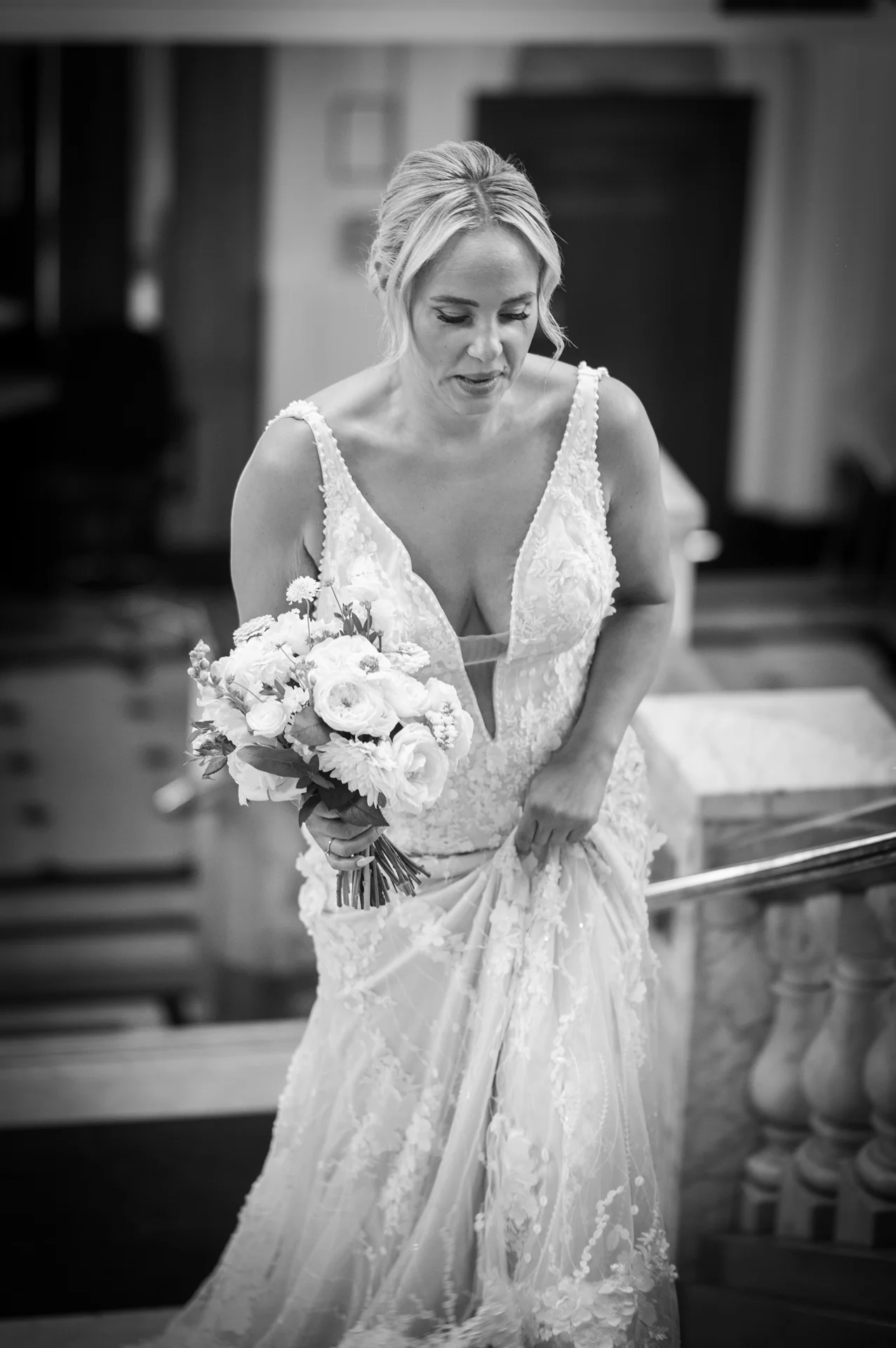 A graceful bride descends a grand staircase in a plunging v-neckline gown with floral appliqués, holding a textured bouquet of white roses.