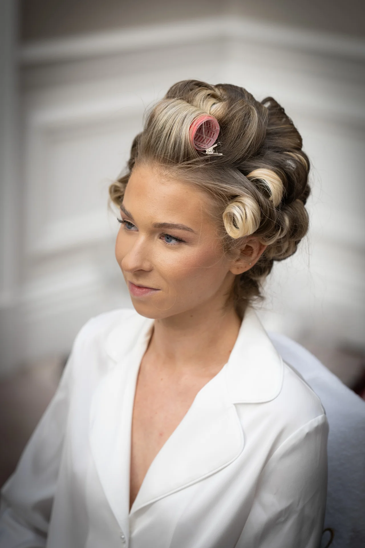 A close-up portrait of a bride getting ready with hair rollers set in her blonde hair while wearing a white robe.