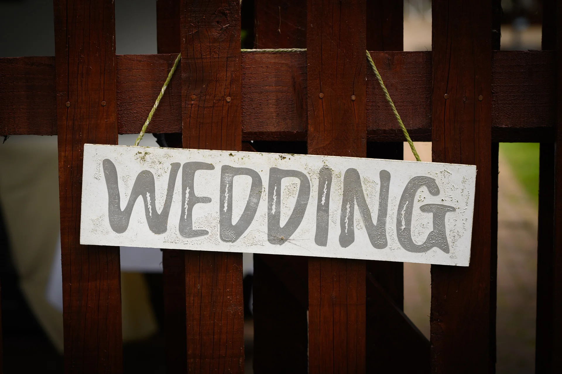 A rustic white wooden sign with the word wedding painted in grey hangs from a dark brown wooden fence.
