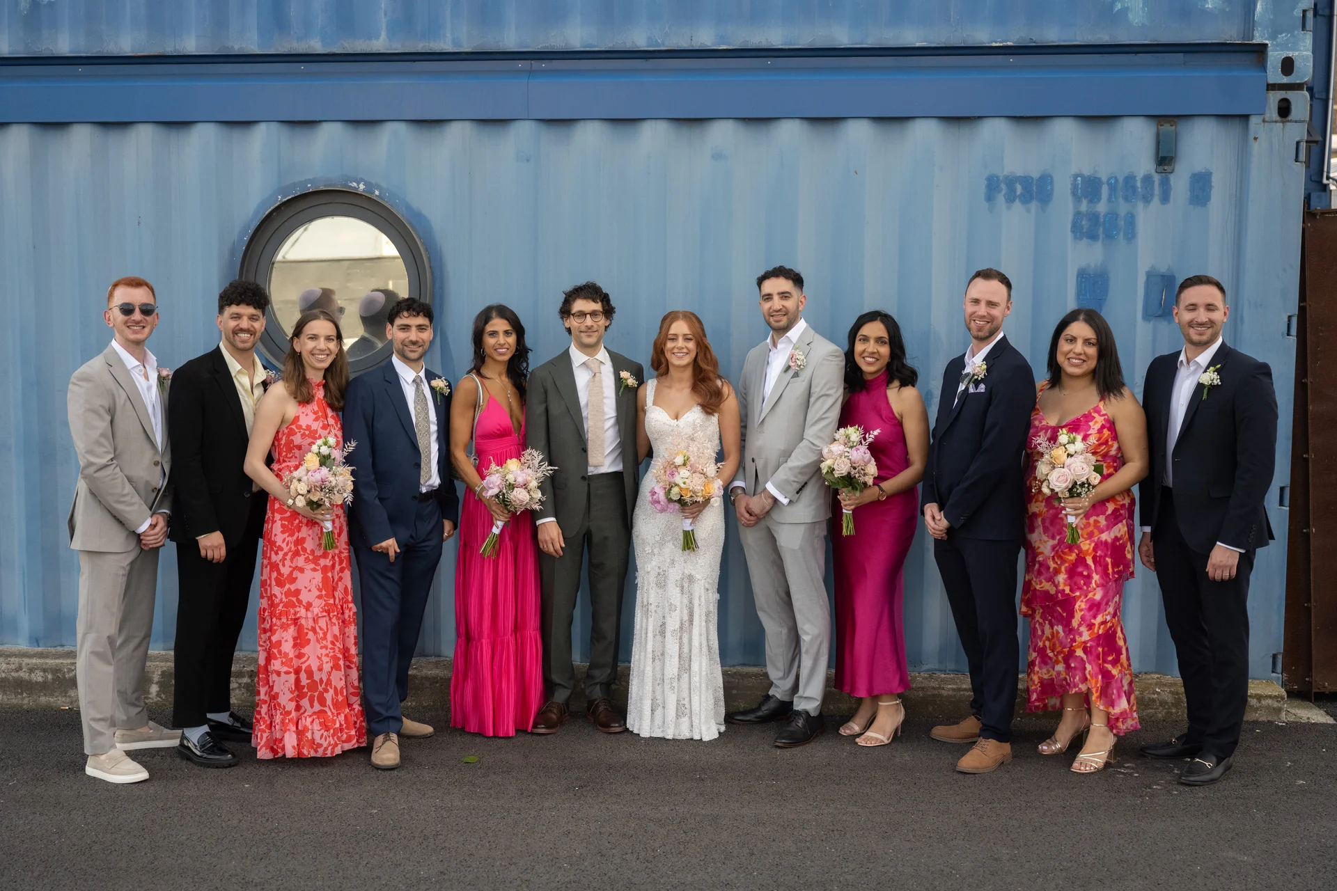A newlywed couple and their wedding party pose together in front of a striking blue industrial container background.