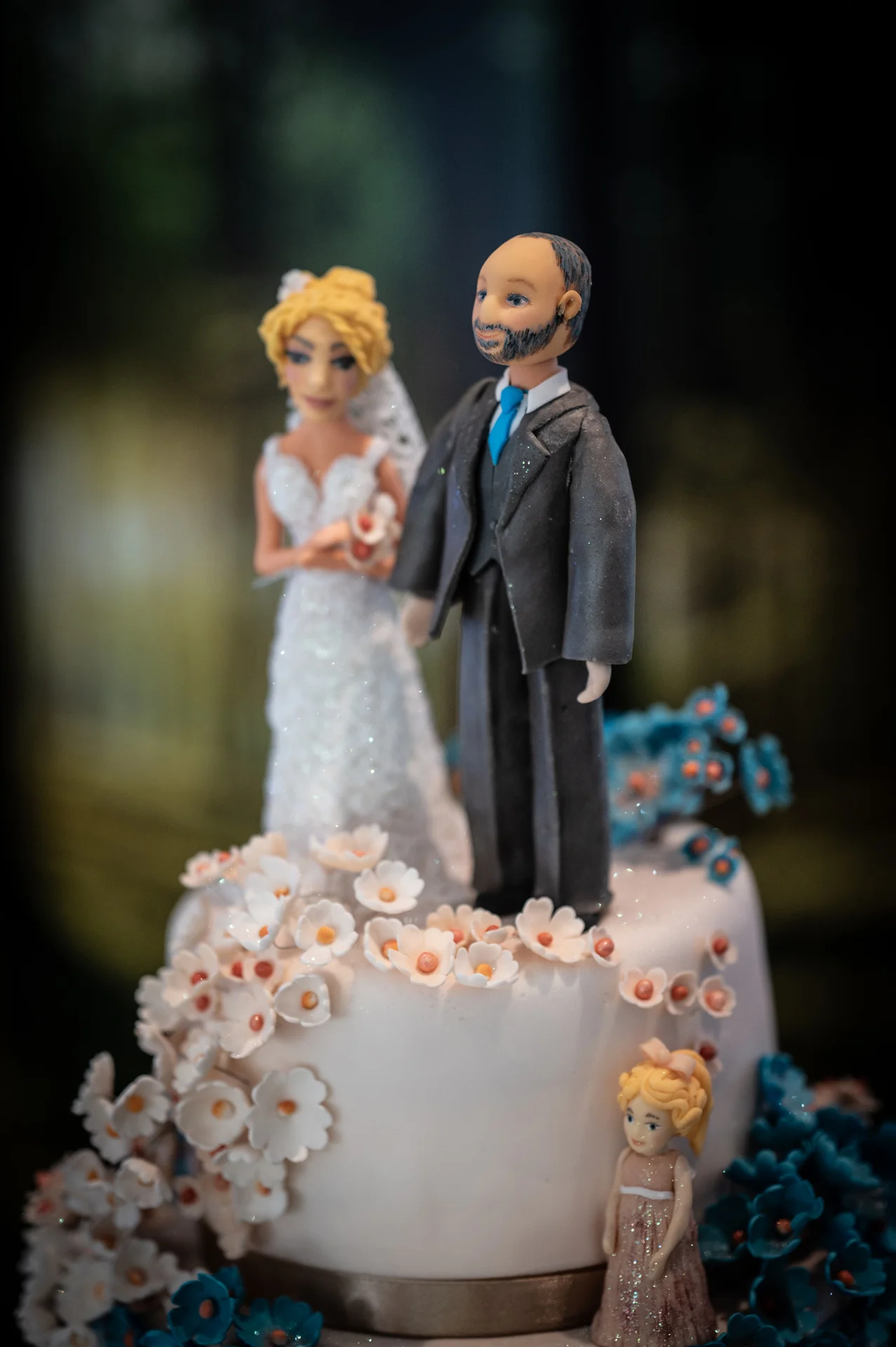 A detailed close-up shot of a white tiered wedding cake featuring intricate sugar flower cascades and a custom bride, groom, and child figurine topper.