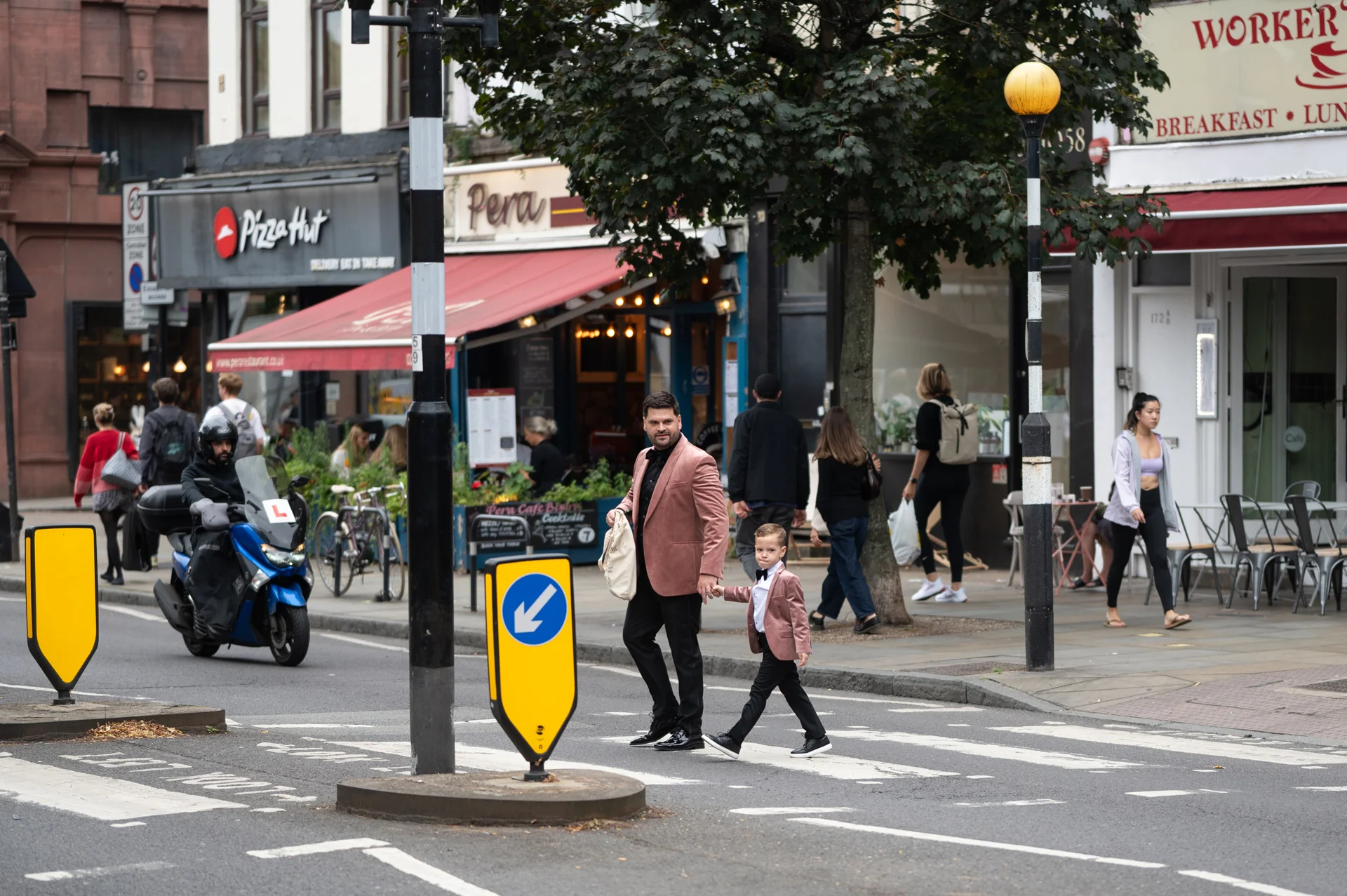 A man and a young boy wearing matching pink blazers walk hand-in-hand across a zebra crossing on a busy city street.