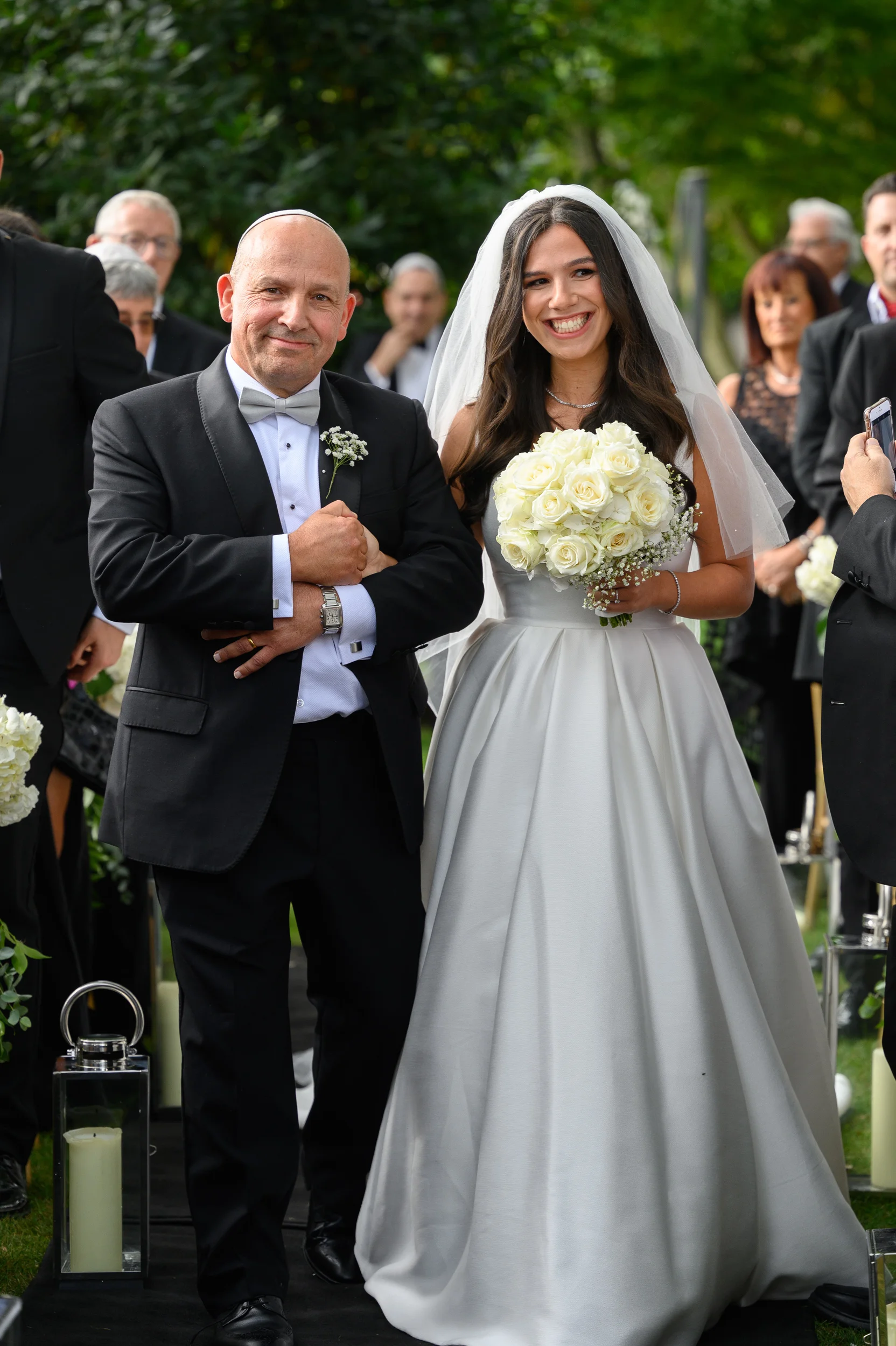 A smiling bride holding a white rose bouquet walks arm-in-arm with her father down the aisle during an outdoor wedding ceremony.