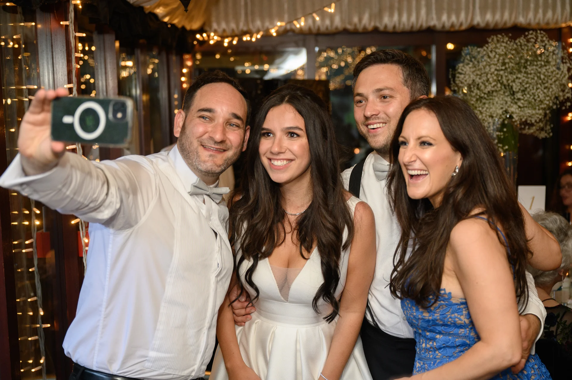 A group of wedding guests smiling and laughing while taking a selfie together at an evening reception illuminated by fairy lights.