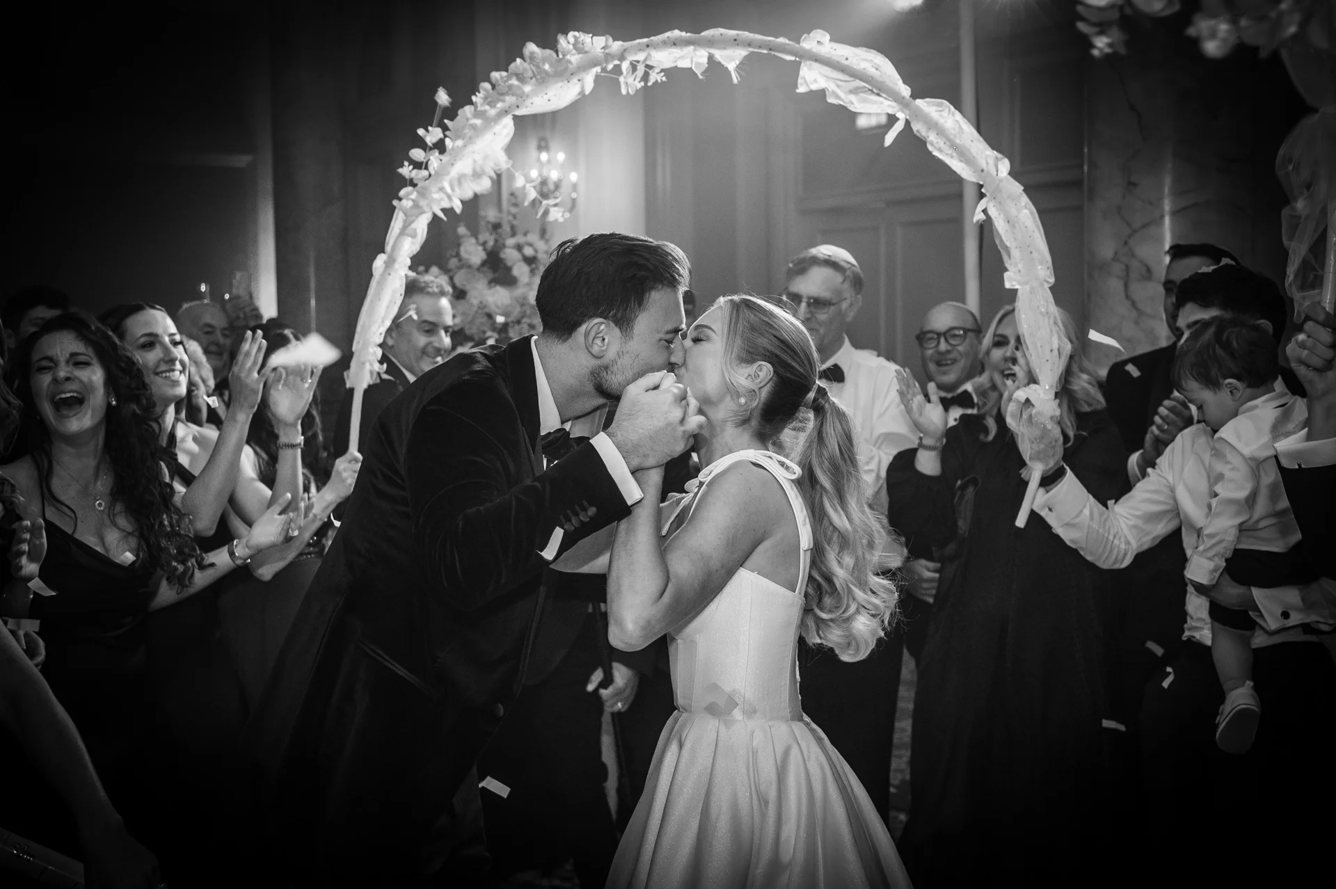 A newlywed couple shares a passionate kiss beneath a flower-adorned arch while celebrating with joyful guests in a grand reception venue.