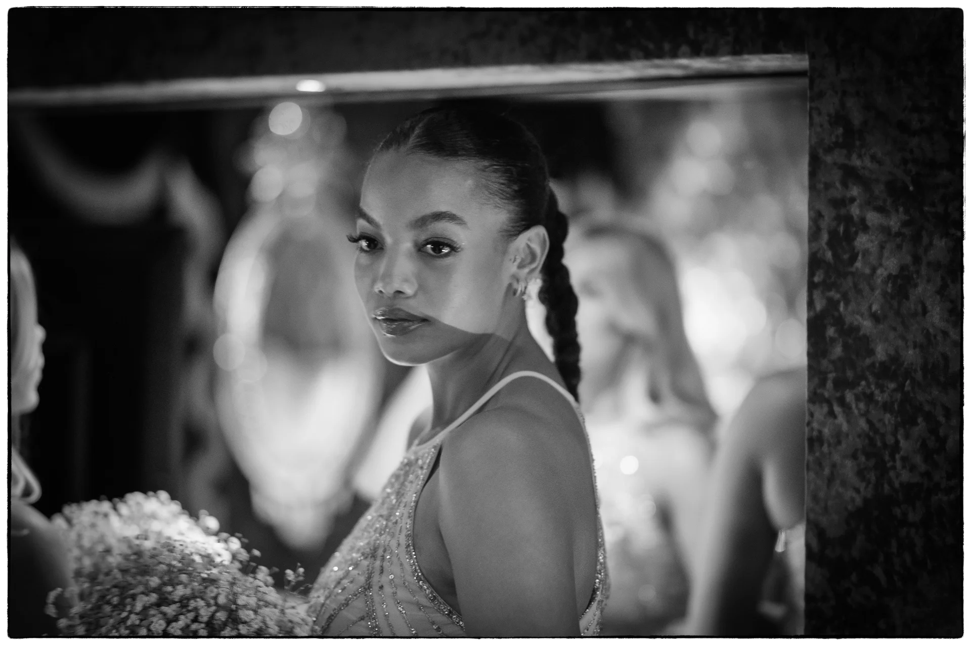 A stunning black and white film portrait of a bride looking over her shoulder into a mirror, wearing an intricately beaded gown with her hair in a sleek braided bun.