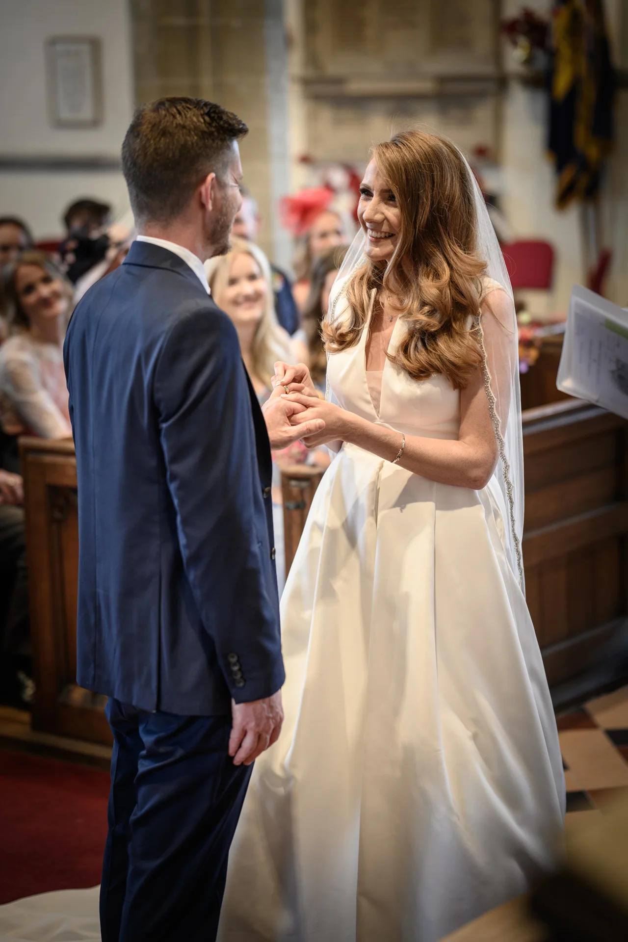 A smiling bride places a wedding ring onto the groom's finger during their church ceremony, wearing a modern satin a-line gown and a delicate embellished veil.