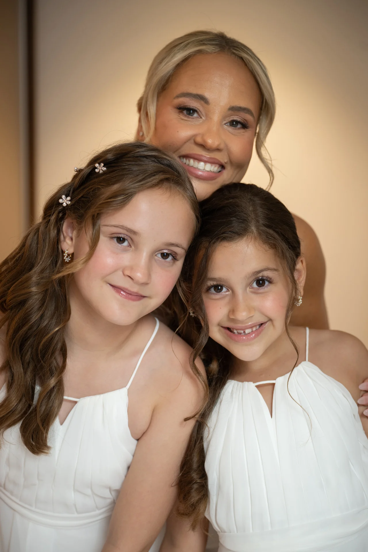 A smiling woman poses closely with two young flower girls wearing white dresses with delicate spaghetti straps and subtle hair accessories.