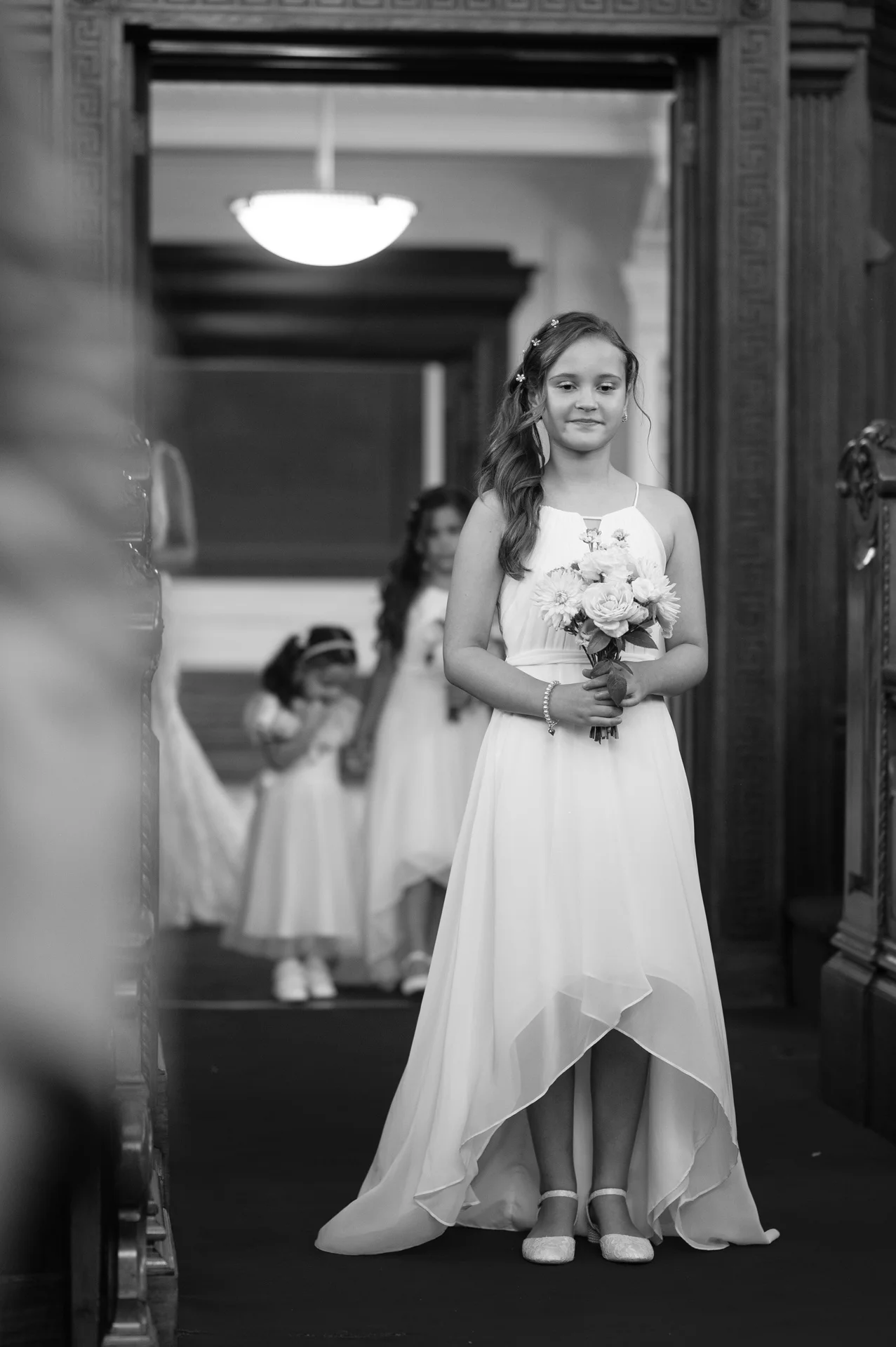 A young flower girl holding a small bouquet walks down the aisle framed by a doorway during a wedding processional.