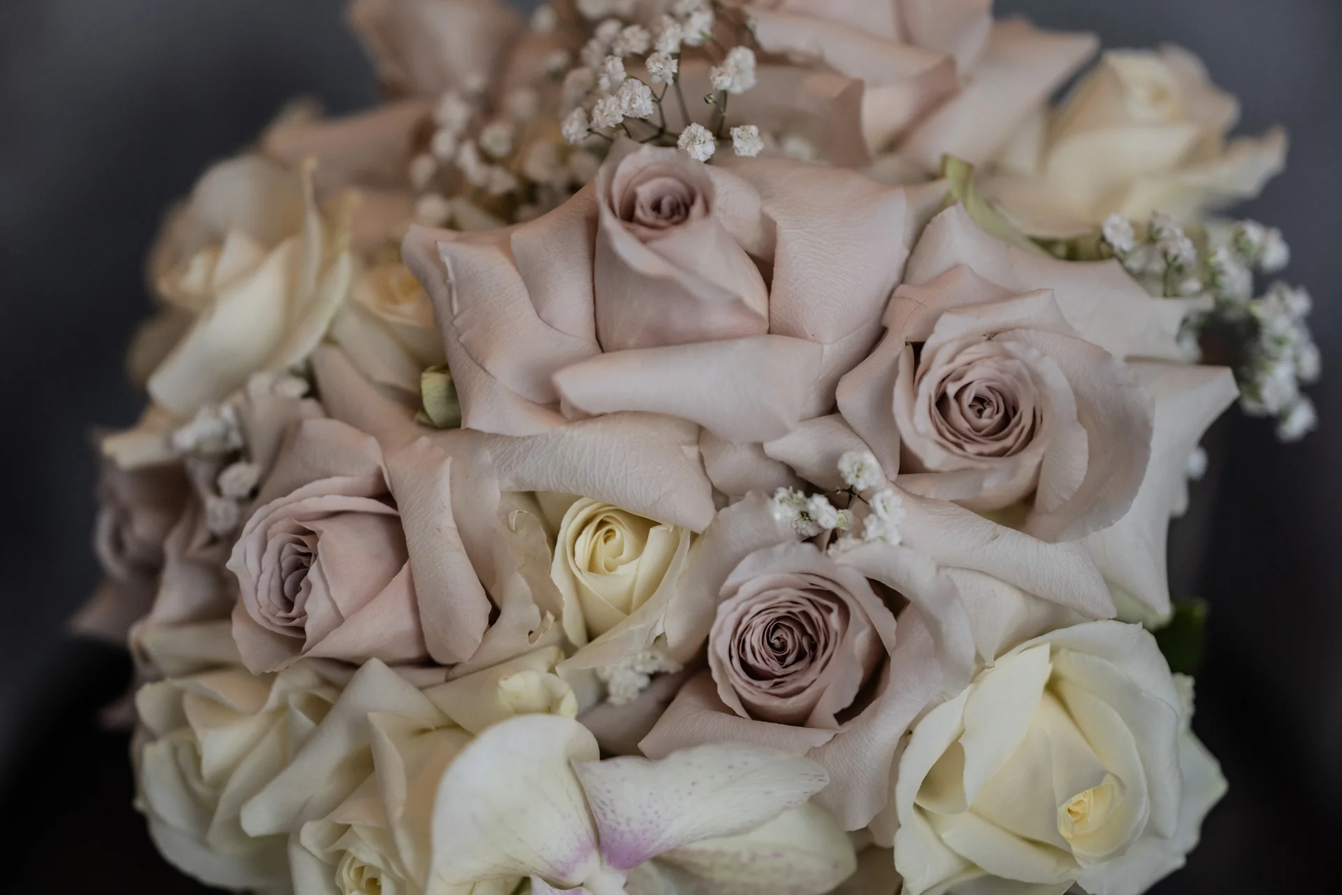 A close-up view of a bridal bouquet featuring pale quicksand roses, white roses, delicate orchids, and sprigs of gypsophila.
