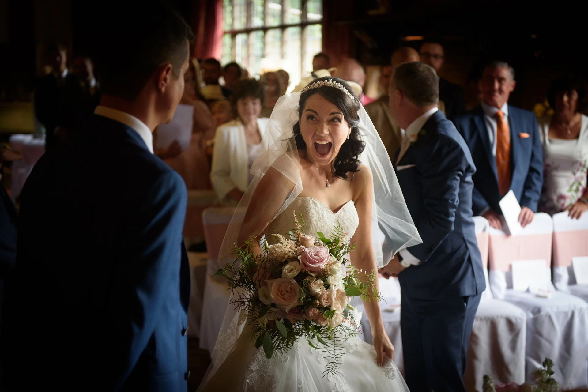 An ecstatic bride laughs joyfully as she walks down the aisle towards the groom, holding a bouquet of roses and greenery during the ceremony.